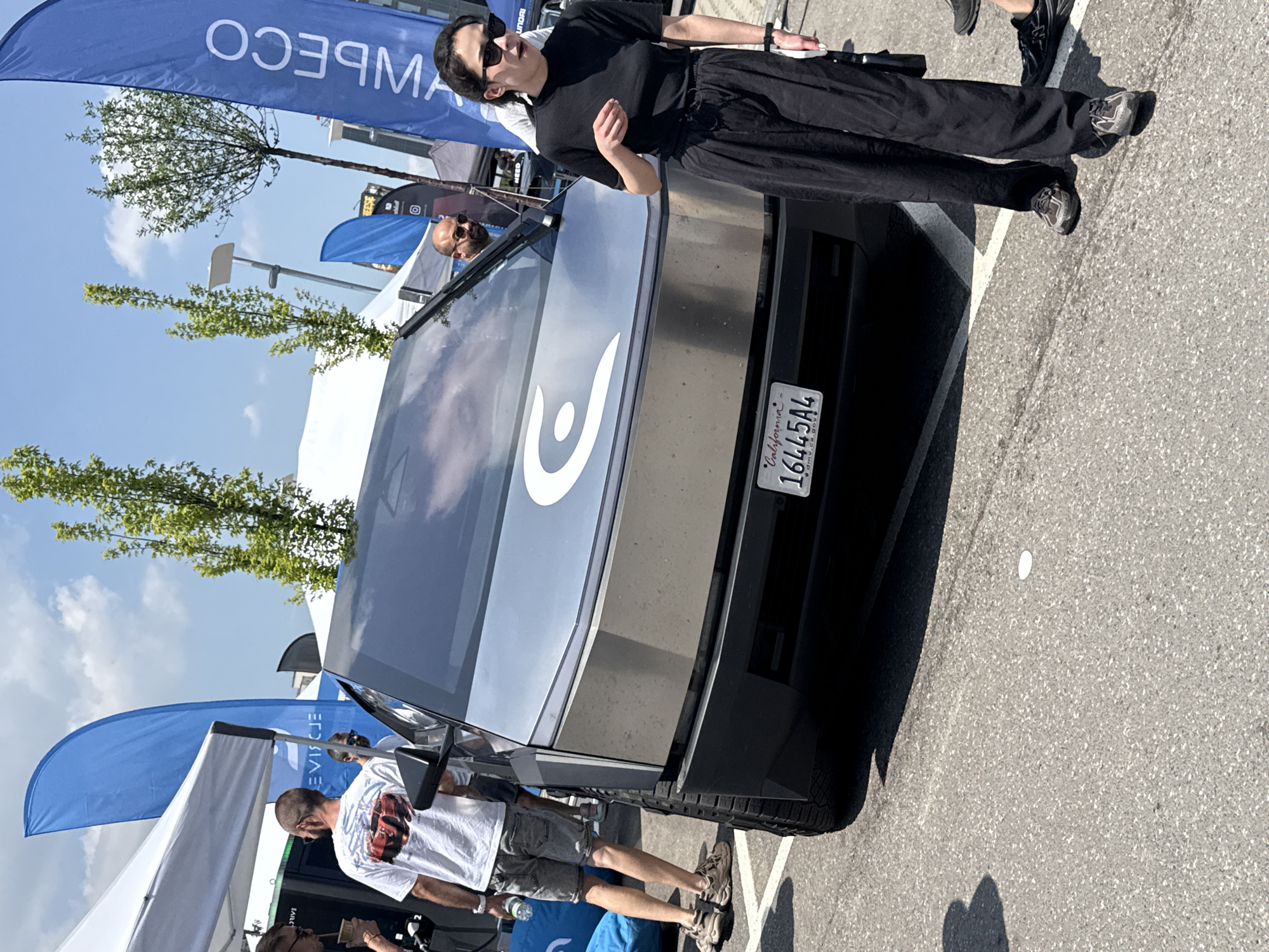 This image captures an outdoor event under a clear blue sky with scattered white clouds, likely a car show or exhibition, during daylight hours.
In the foreground, a metallic silver Tesla Cybertruck is parked, angled slightly towards the right. A large white 'C' logo is prominently displayed on its front. The vehicle has a visible California license plate, "16445A4", which includes the text "dmv.ca.gov". This indicates the event is taking place in California, USA, contradicting the suggestion of Sofia, Bulgaria. The Cybertruck's tires are large and knobby, suggesting off-road capability.
To the right of the Cybertruck, an adult woman stands, facing left towards the vehicle. She wears a short-sleeved black top, loose-fitting black wide-leg pants, black athletic shoes (resembling New Balance), and black sunglasses. Her left hand is slightly raised, and her right hand holds a dark object, possibly a phone or a small purse. She appears to be observing or in conversation.
Inside the Cybertruck, visible through the windshield, are two adult men. The man in the driver's seat has a bald or shaved head and is wearing sunglasses. Another man, also with a bald or shaved head and wearing sunglasses, is visible through the windshield further to the right.
To the left of the Cybertruck, partially visible behind a white canopy tent, an adult man stands with his back mostly to the camera. He wears a white t-shirt featuring a red sports car graphic on the back, grey shorts, and light-colored athletic shoes. He holds a clear plastic water bottle in his right hand. A light blue beanbag-like object rests on the ground near his feet.
Further into the background, several blue feather flags or banners are visible. One on the far left, behind the man in the white shirt, shows partial text "EVIJE" (reversed). Another prominent blue banner on the far right displays "ОЭЯМ" (reversed), which would read as "MЭCO" or similar from the front, possibly part of a brand name like "AMBECO" or "ECOMBE". White canopy tents are present on both sides of the scene, and several young, leafy trees are scattered in the mid-ground. The ground is paved with asphalt, showing white parking lines and a small white disc (possibly a discarded lid or paper) in the lower foreground.