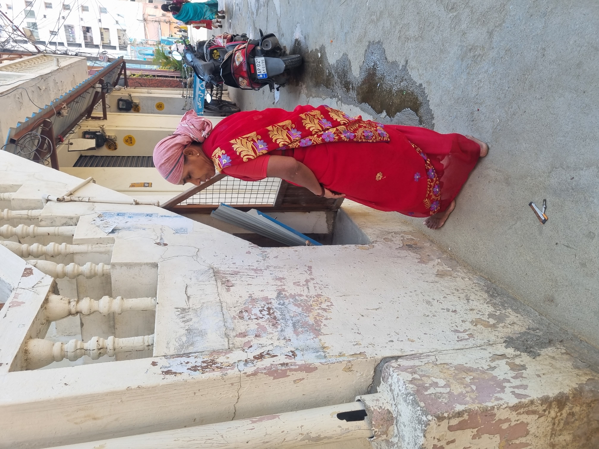 The image depicts a street scene in Pedda Kammavaripalli, India, under bright daylight. In the foreground, an adult female with a medium-dark complexion stands barefoot on a light grey paved surface. She wears a red sari with extensive gold and purple floral embroidery, a matching red blouse, and a light pink cloth wrapped around her head. She faces towards a cream-colored building wall on her left, slightly turned from the camera, with her right arm bent towards her side.
The adjacent building wall exhibits significant weathering, with extensive peeling cream paint revealing patches of pinkish and grey undercoat. A portion of a white concrete staircase with balusters is visible above the wall, also showing signs of wear. A small, recessed opening, partially covered by a metal grate or shutter, is present in the wall below the stairs. A small, dark-colored object, possibly a utility knife, lies on the paved ground near the woman's feet. Several irregular dark wet patches are visible on the pavement.
In the midground, a red Honda Activa scooter (license plate AP 02 AY 6096) is parked, facing away from the camera. Behind it, part of a blue-painted cart or rickshaw is discernible. The buildings lining the street feature corrugated metal awnings, under which exposed electrical wiring bundles are visible. Multiple utility meters or junction boxes with yellow labels (e.g., "14405," "14406," "14412") are affixed to the building exteriors. Further down the street, another female in a turquoise sari walks with a child, both facing away. Tall, multi-story buildings are visible in the distant background.