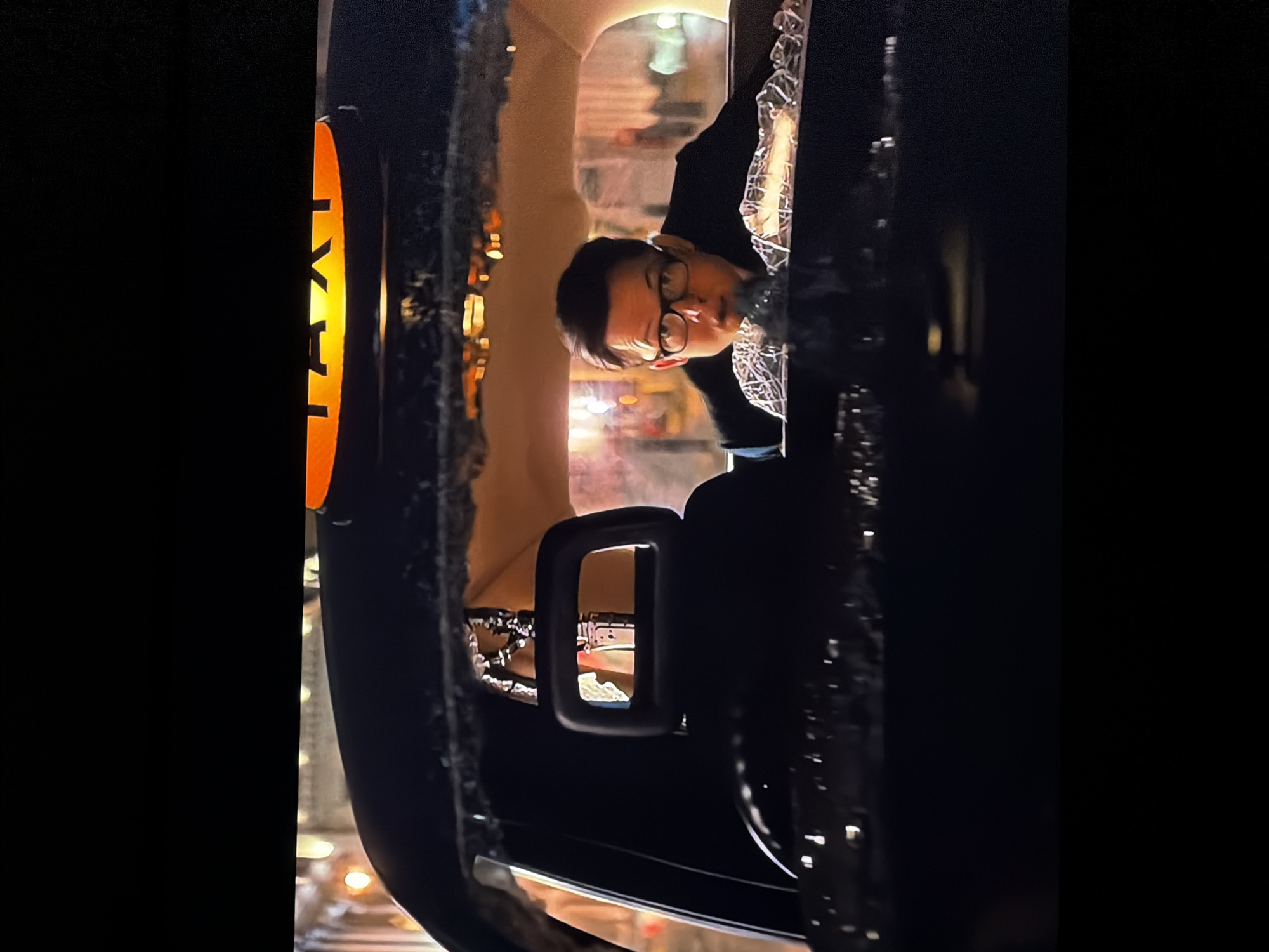 A nocturnal, candid shot features a person in the interior of a taxi in Sofiivska Borshchahivka, Ukraine. The image is framed from an exterior perspective, looking into the vehicle. A bright, illuminated "TAXI" sign, rendered in black text against a yellow-orange background, is prominently visible on the left side of the vehicle's roof, signifying its purpose.
Inside the dark-colored taxi, a person with short, dark hair and glasses is visible, appearing to look forward and slightly upwards from their seated position. They wear a dark-colored jacket or hoodie, and their expression is neutral. The interior of the taxi, particularly the ceiling and trim, has a textured, possibly fabric or vinyl, finish in a light beige or off-white color. Reflections of distant lights, blurred and indistinct, are visible through the vehicle's windows, suggesting a city environment at night. A part of what appears to be a rearview mirror or an interior handle is also visible within the cabin. The overall atmosphere is one of urban transit during evening hours.