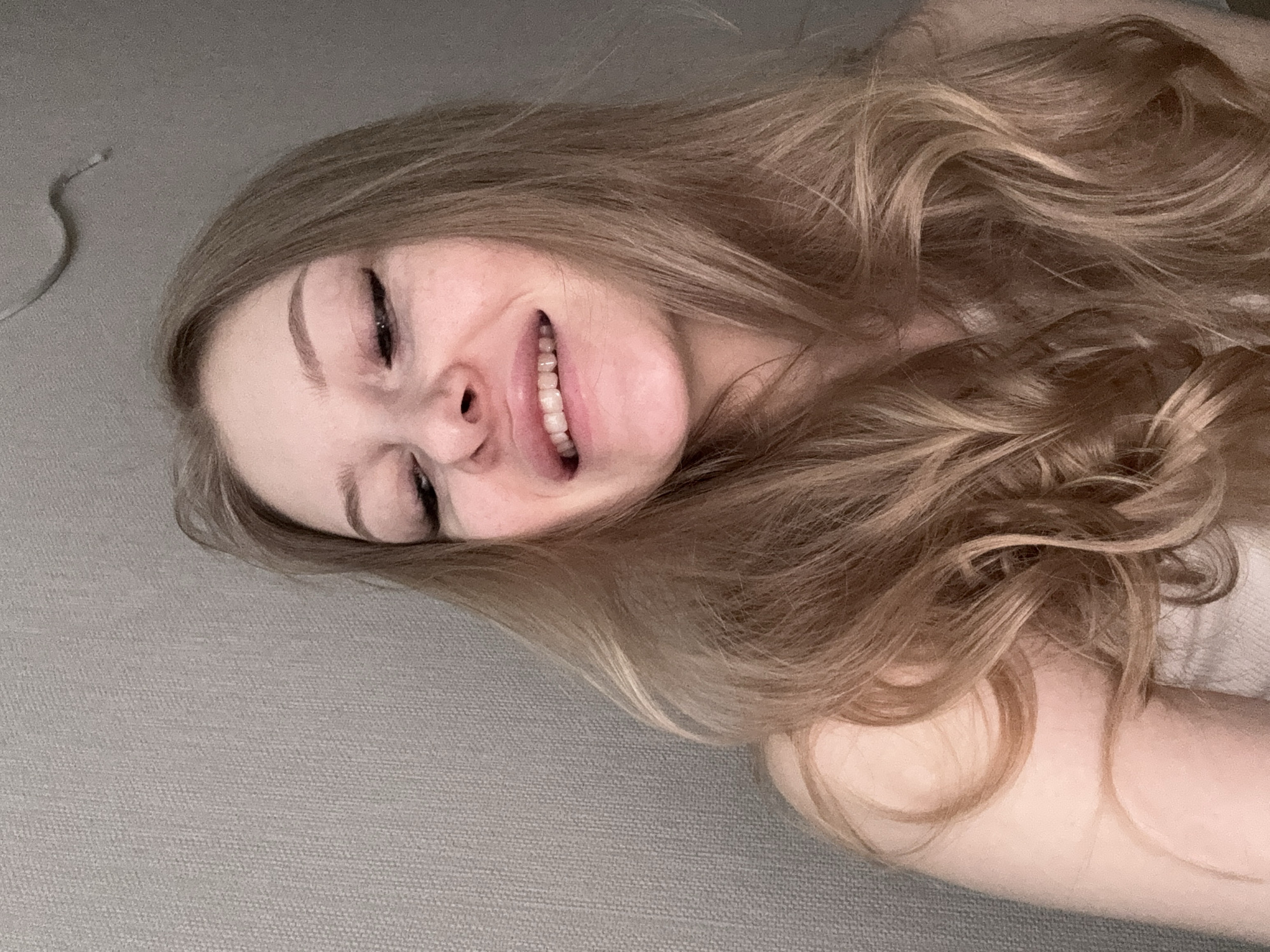 A woman with long, wavy, light brown hair is lying on a grey surface, possibly a bed or couch. She is smiling broadly, with her eyes partially closed. The background is a plain grey texture. The setting does not appear to be outdoors or in a public location in Perm, Russia.