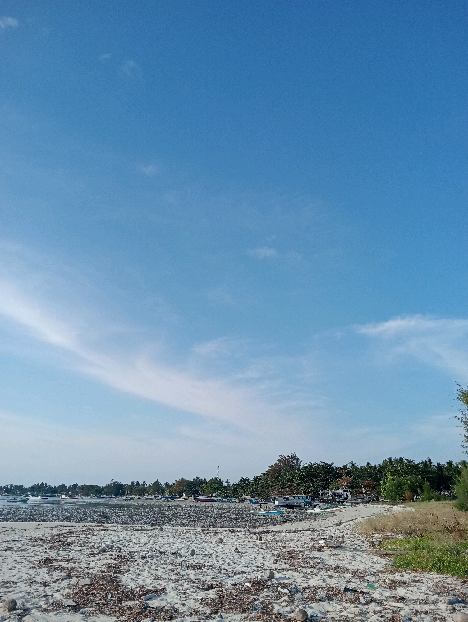 The image shows a beach in Pagerungan Kecil, Indonesia. The foreground is a sandy beach with scattered debris. Behind the sand, there is a rocky shoreline with several boats anchored. The middle ground features a line of trees and vegetation along the coast. In the distance, more trees and structures are visible. The sky is clear blue with some wispy clouds.
