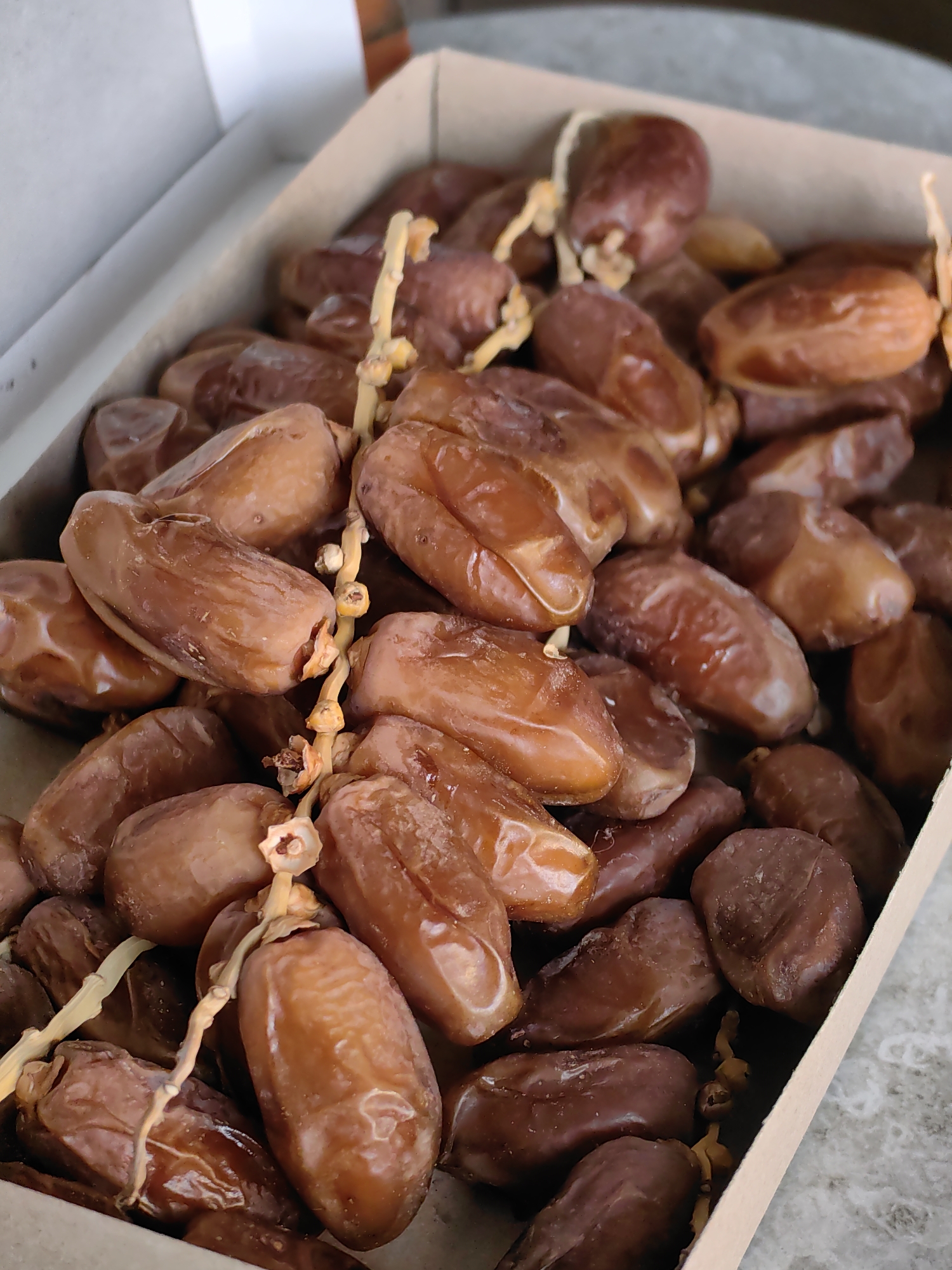A close-up shot shows a cardboard box filled with fresh dates. The dates are a rich brown color and some are still attached to their stems. The box is positioned on a textured, gray surface. The background is blurred, with hints of a brick wall and a white surface. The image is taken from a slightly elevated angle, looking down into the box. There are no people or animals visible in the image.