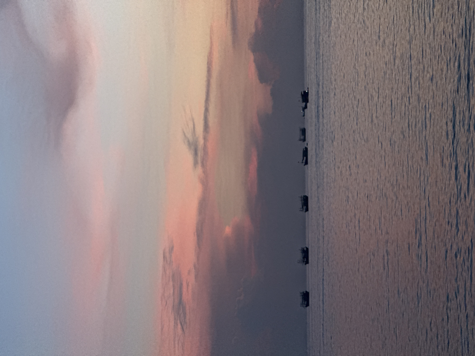 The image depicts a tranquil seascape at dusk or dawn, with a pastel sky blending shades of pale blue, pink, and soft orange from the setting or rising sun. Several small fishing boats are silhouetted against the horizon, scattered across the calm, rippling water. The water reflects the muted colors of the sky. The scene is likely located off the coast of Lý Sơn Special Administrative Region, Vietnam, a region known for its fishing villages and beautiful coastal landscapes. There are no people visible in the image.