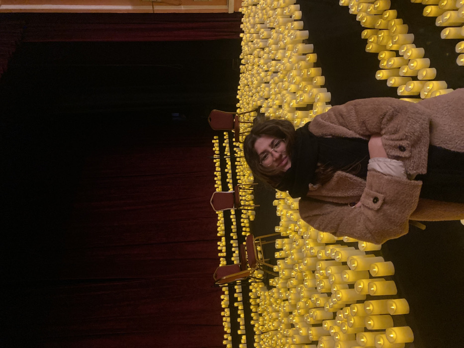 An indoor scene depicts a dimly lit theater or concert hall in Sofia, Bulgaria, illuminated primarily by hundreds of small, glowing LED candles. A female individual, appearing to be an adult with medium-length dark hair and glasses, stands in the foreground, slightly off-center to the left. She wears a light brown, possibly faux fur, coat and a black scarf. Her arms are crossed, and she is smiling faintly with her head tilted to her left. The floor around her and extending into the midground is covered densely with rows of cylindrical LED candles, emitting a warm yellow light. In the background, three empty, dark red chairs with gold-colored frames are arranged on a raised stage area, also surrounded by the candles. Behind the chairs, large, dark red velvet stage curtains extend across the width of the frame, indicating a performance venue. The upper portion of the background is dark, suggesting the stage ceiling.