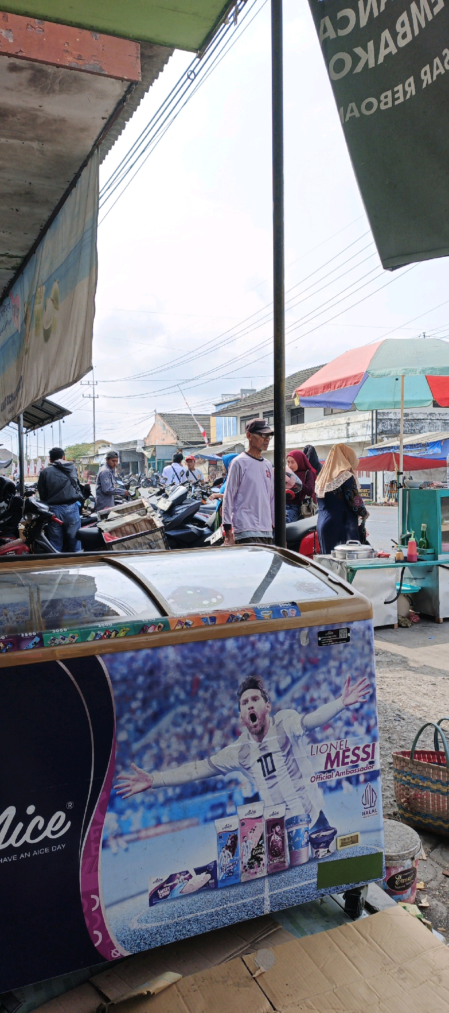 The image depicts an outdoor market scene in Kasiyan, Indonesia, characterized by a bright, overcast sky and the presence of various stalls and vendors. In the foreground, a large freezer chest is prominently displayed, featuring a vibrant advertisement of Lionel Messi in his football kit. The freezer is adorned with images of ice cream products and branding, including "Aice" and "HALAL INDONESIA".
The market area is bustling with activity. Several individuals are seen in the mid-ground, including men and women dressed in casual attire. Some are interacting with each other, while others appear to be engaged in shopping or work. Motorcycles are parked closely together, suggesting a common mode of transportation in the area.
Overhead, power lines crisscross the sky, indicating the presence of urban infrastructure. Several market stalls with awnings and umbrellas are visible, offering shelter from the elements. The background reveals a mix of residential buildings and commercial structures, typical of a developing urban environment in Indonesia. The overall atmosphere suggests a lively marketplace on a typical day.