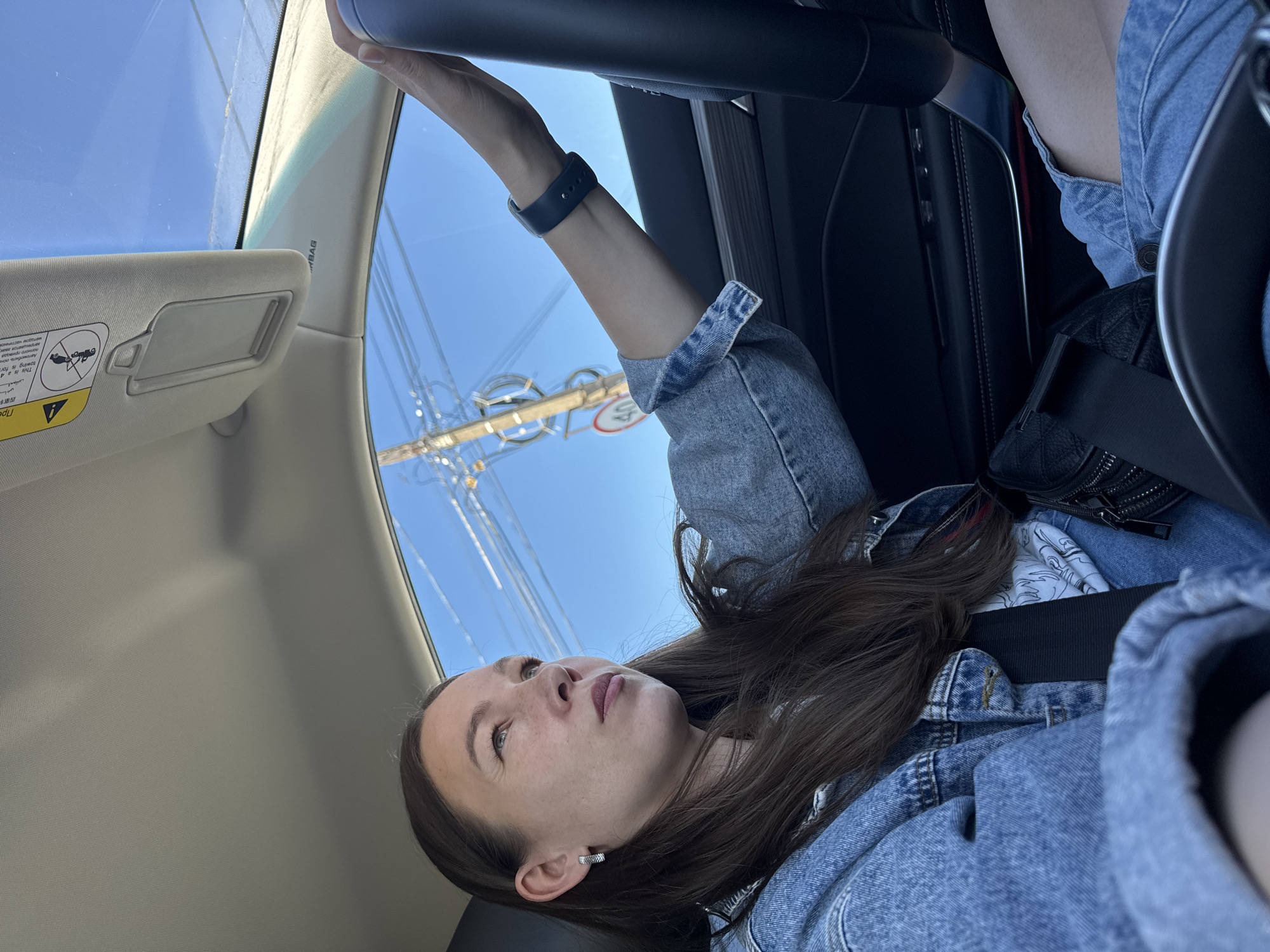 A woman is sitting in the driver's seat of a car, looking out the window. She is wearing a blue denim jacket and a black smartwatch. The car is parked on a street in Chaikovsky, Russia, with power lines visible through the windshield. The sun visor is down on the driver's side. The woman is wearing a seatbelt.