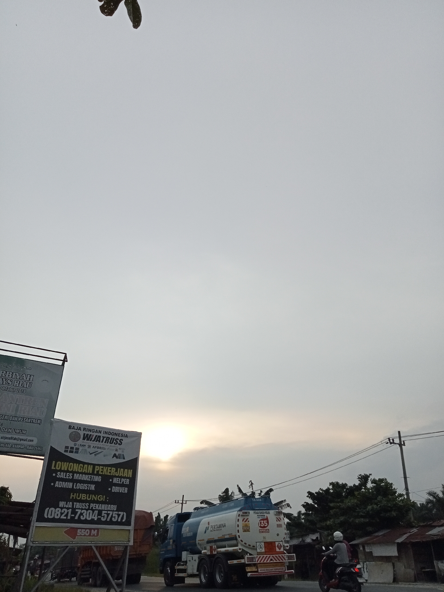 The image depicts a roadside scene in Kubang Jaya, Indonesia, under a hazy, overcast sky with the sun partially visible through the clouds, suggesting it is late afternoon or early morning. In the foreground, a large billboard displays "BAJA RINGAN INDONESIA WIJATRUSS" and "LOWONGAN PEKERJAAN" (Job Vacancy) with listings for Sales Marketing, Admin Logistik, Helper, and Driver, along with contact information. Another partially visible billboard to the left appears to be related to an educational institution, possibly mentioning "PESANTREN" (Islamic boarding school) and "RIAU".
To the right of the billboards, a Pertamina fuel tanker truck is parked on the road. The truck has the Pertamina logo and is labeled with information such as "PREMIUM", "SUPER", and a safety warning with the number "35". A motorcycle carrying two individuals, one wearing a white helmet, is traveling on the road in front of the fuel truck. In the background, there are trees and several buildings with corrugated metal roofs. The overall impression is of a commercial and transportation hub along a road in a semi-urban or rural area.