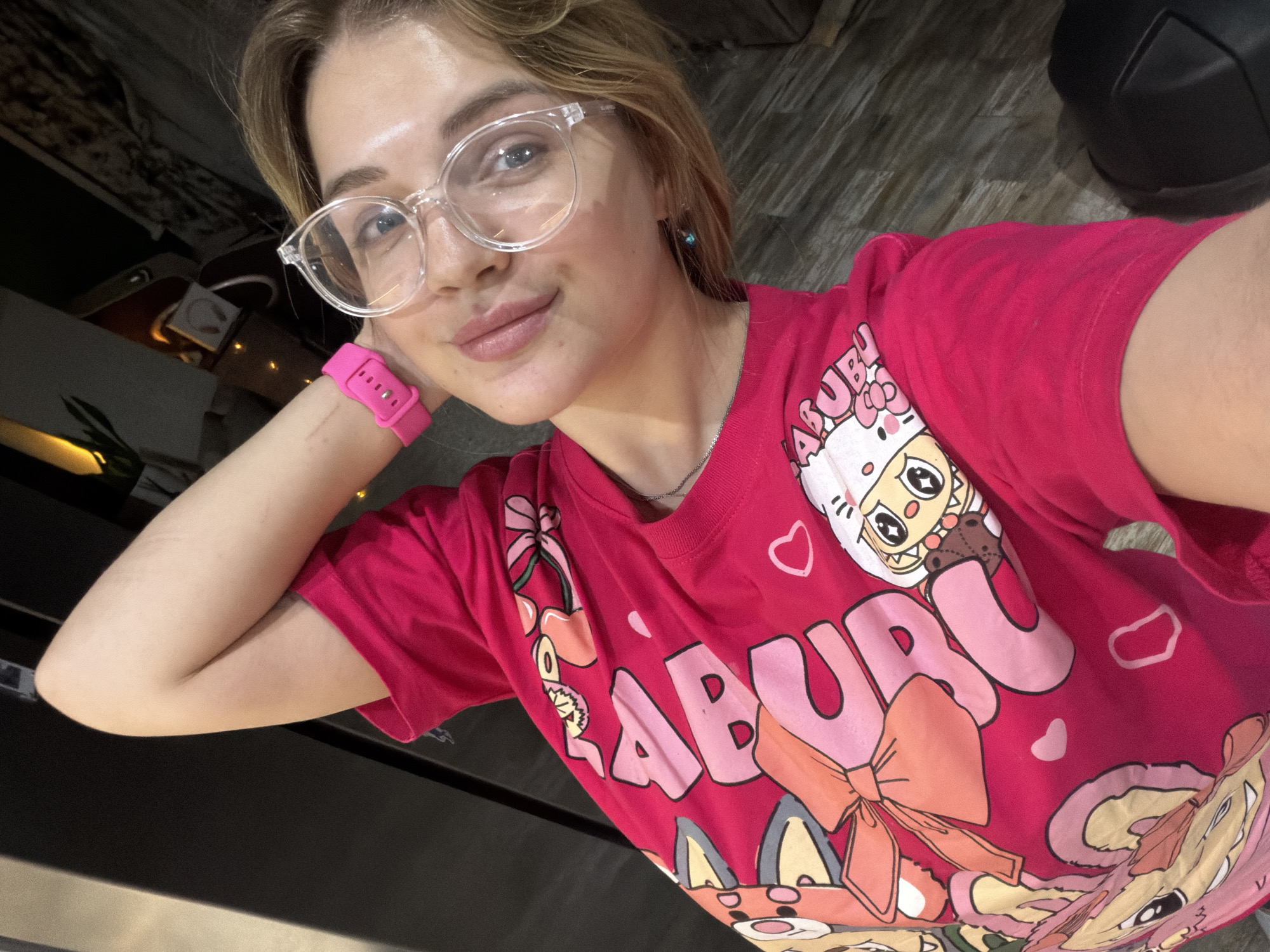 A young woman is captured in a close-up selfie, taken indoors in Большая Шильна, Russia. She has light skin and blonde or light brown hair, and is wearing clear-framed glasses. Her expression is relaxed and pleasant, with a subtle smile. A vibrant pink silicone watch is prominent on her left wrist, which is bent, with her hand gently supporting her head or chin. She also wears a thin silver chain necklace.
The woman is wearing a bright pink t-shirt adorned with various cartoon characters and large pink text. The word "BABURU" is clearly visible in stylized pink letters on the front of the shirt. Other designs include a character resembling Hello Kitty with a bow, additional cartoon animal-like figures (such as a fox, cat, and squirrel), multiple hearts, and a design featuring cherries.
The background is softly blurred, showing dark, indistinct shapes that suggest furniture or structural elements of an indoor setting. Hints of a patterned floor, possibly wood or tile, are discernible, along with some artificial lighting in the deeper background. The overall lighting is dim, indicating an indoor environment, potentially in the evening or a room with low natural light. The woman is looking directly at the camera.