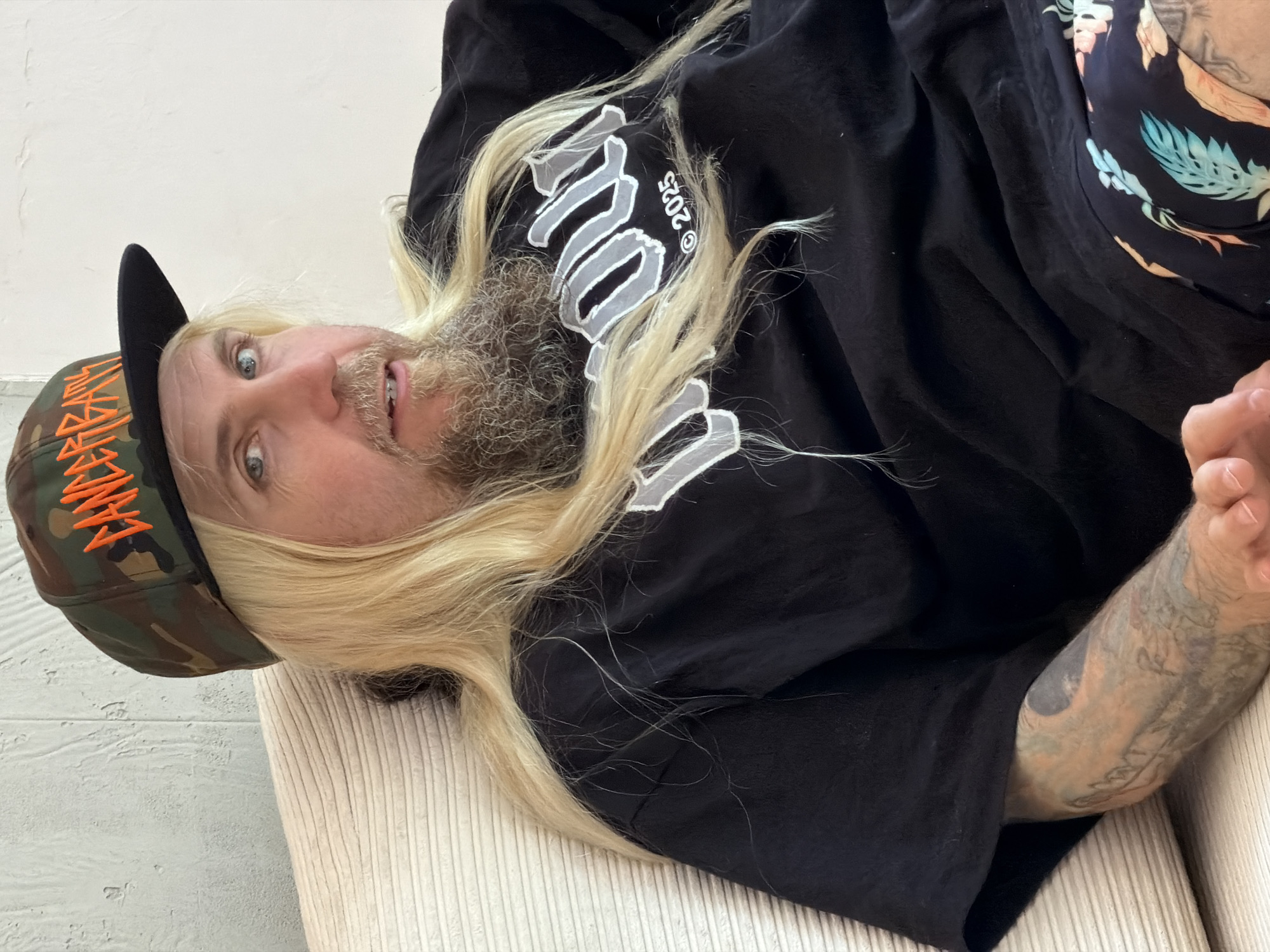 A person with long blonde hair and a beard lies on a light-colored, ribbed surface, possibly a couch or chair. They are wearing a camouflage baseball cap with orange lettering, a black t-shirt with white lettering and the year "2025," and floral shorts. The person has tattoos on their arm. The background appears to be a plain white wall. The image provides no information to determine if the location is Budva, Montenegro.