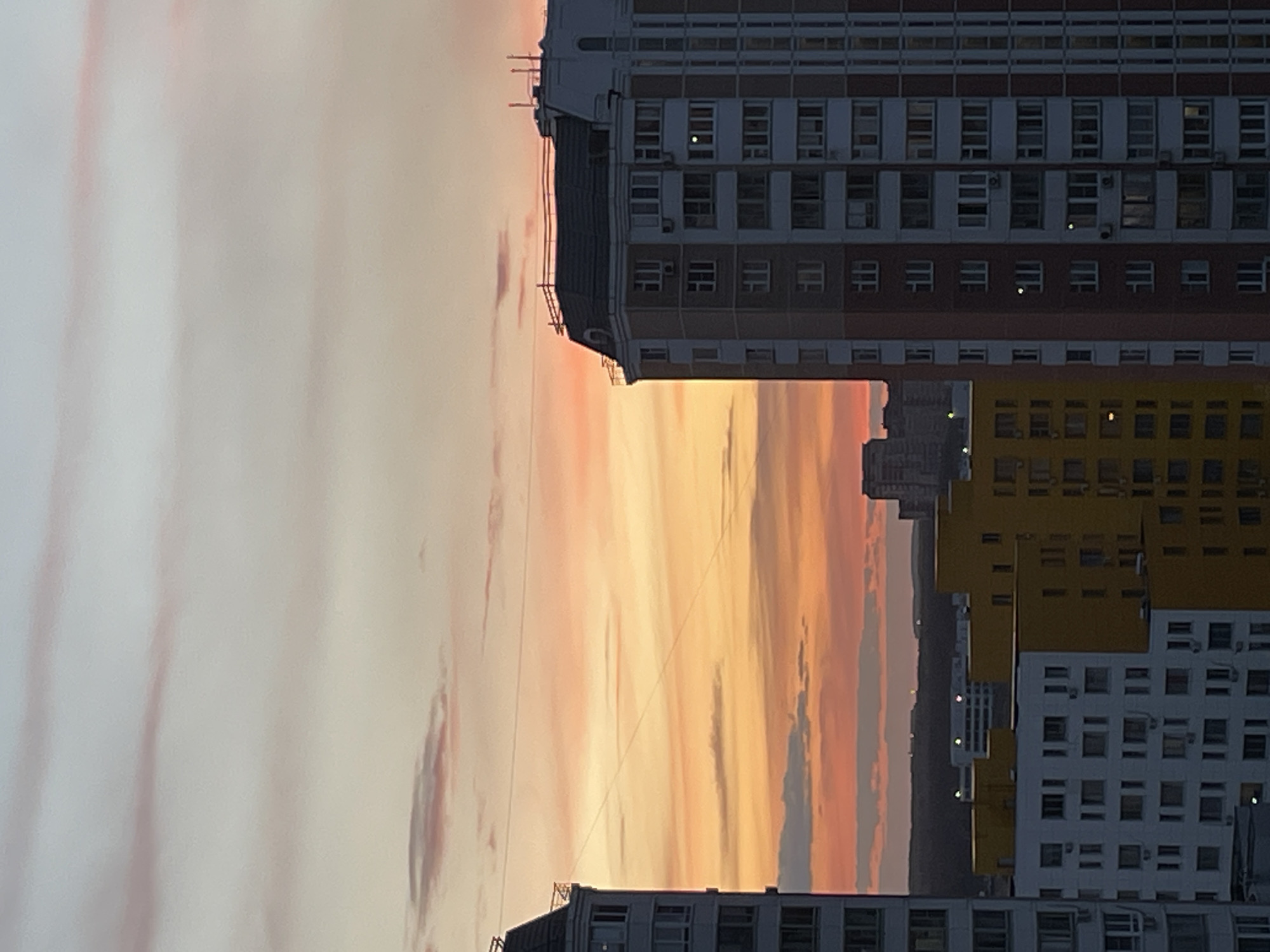 The image shows a view of buildings in Moskovskiy, Russia, at sunset. The sky is filled with pink, orange, and gray hues. In the foreground are several high-rise apartment buildings of varying heights and colors, including white, yellow, and brown. The buildings have multiple rows of windows, with some windows illuminated. In the background, silhouetted buildings are visible. Power lines stretch across the sky. The image has been rotated, placing the skyline on a vertical axis.