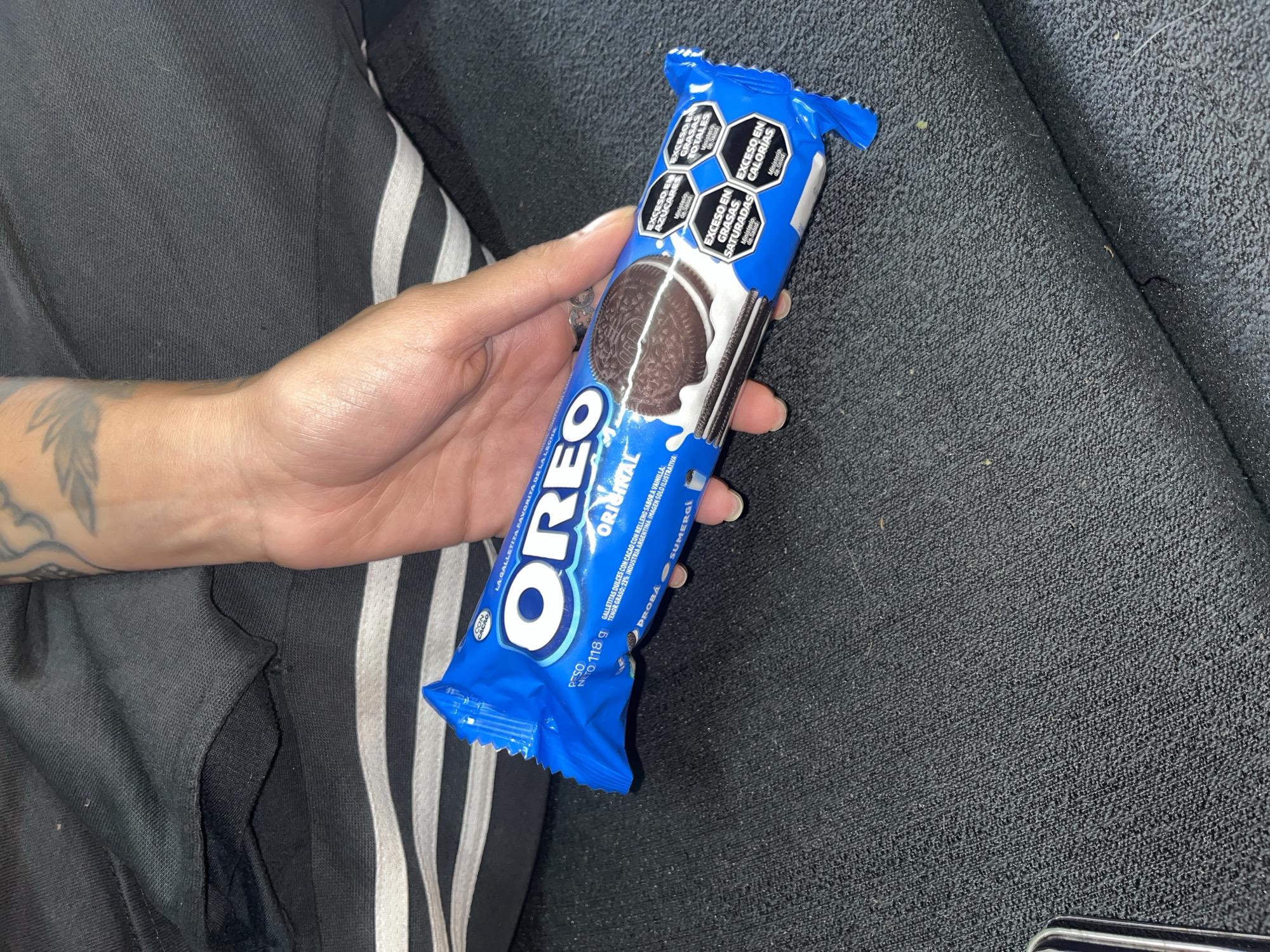 A human hand holds a blue, unopened package of Oreo Original cookies. The package has warning labels indicating excessive amounts of sugar, saturated fats, total fats, and calories. The background shows black fabric and black athletic pants with white stripes. The packaging indicates the product is from Argentina. The location of Llavallol cannot be determined from the image.