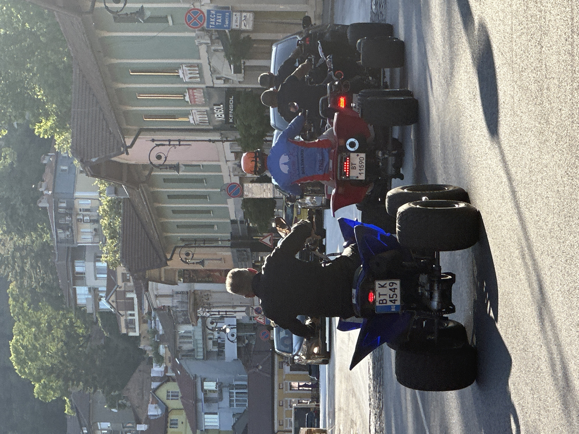 The image shows a street scene in Veliko Tarnovo, Bulgaria, with several people riding ATVs. In the foreground, a person in a black jacket rides a blue ATV with the license plate BT K 4549. To the right, a person in a blue shirt and orange helmet rides a red ATV with the license plate BT 11590. Behind them, two more individuals are riding ATVs, and a dark-colored car is visible. The background features buildings typical of a European city, with a hilly terrain and trees. The scene appears to be set during the daytime, with sunlight casting shadows on the asphalt.