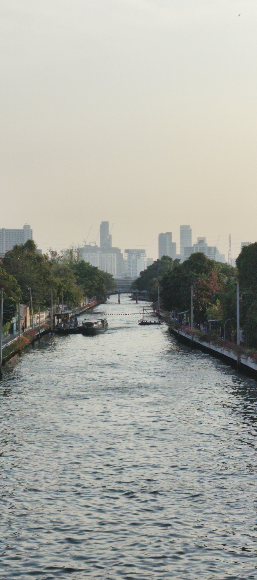 The image presents a vertical view of an urban canal in Bangkok, Thailand, extending into the distance under a hazy sky. The canal, filled with rippled water, dominates the lower and central parts of the frame, leading the eye towards a distant city skyline.
Both banks of the canal are lined with dense green foliage from trees and some low-rise buildings or structures. On the left bank, closer to the viewer, there are a few visible figures near what appears to be a small docking area. A dark-hulled boat, possibly a passenger vessel, is seen navigating the canal in the mid-ground, moving away from the viewer. On the right bank, further back, another floating structure or platform is moored. Two bridges are visible in the distance, spanning the canal.
The background is characterized by a blurred, pale yellow-white sky that merges with a distant, hazy cityscape. Numerous high-rise buildings are discernible, some with cranes indicating ongoing construction, against the soft, diffused light. A small bird is visible in the upper right quadrant of the sky. The time of day appears to be daytime, possibly late morning or afternoon, due to the soft, even lighting and lack of harsh shadows. The overall atmosphere is calm and mundane. No discernible text is visible.