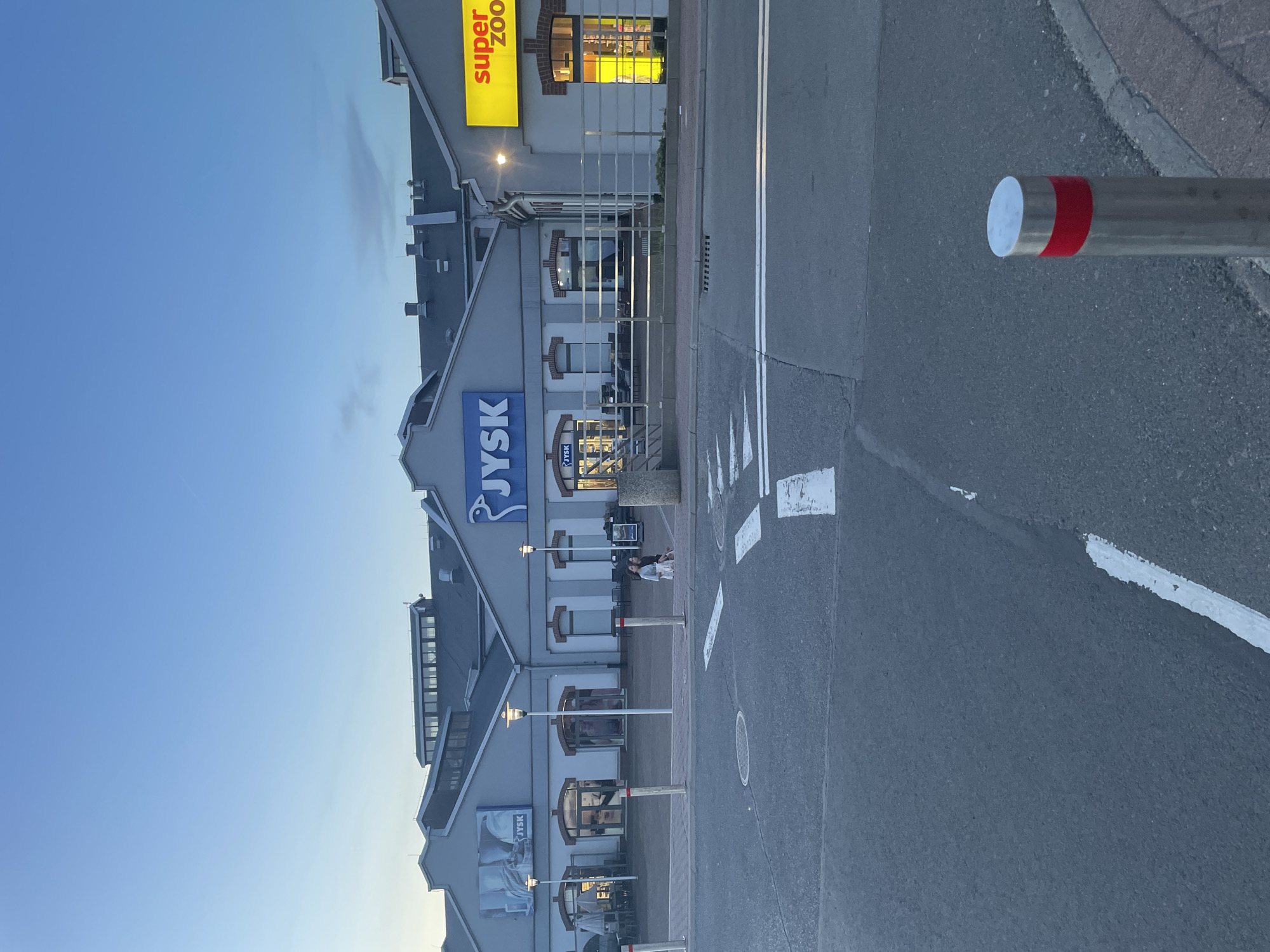 The image shows a street scene in Unknown, Unknown, featuring a building with a JYSK store sign and a Super Zoo store sign. The building is multi-storied with multiple windows and arched decorations above the windows. The sky is a clear blue with a slight gradation towards the horizon. The street is asphalt with painted white lines and chevrons. A grey metal bollard with a red stripe near the top is visible on the right side of the frame. Two people are sitting near the stores in the background. Street lights line the street.