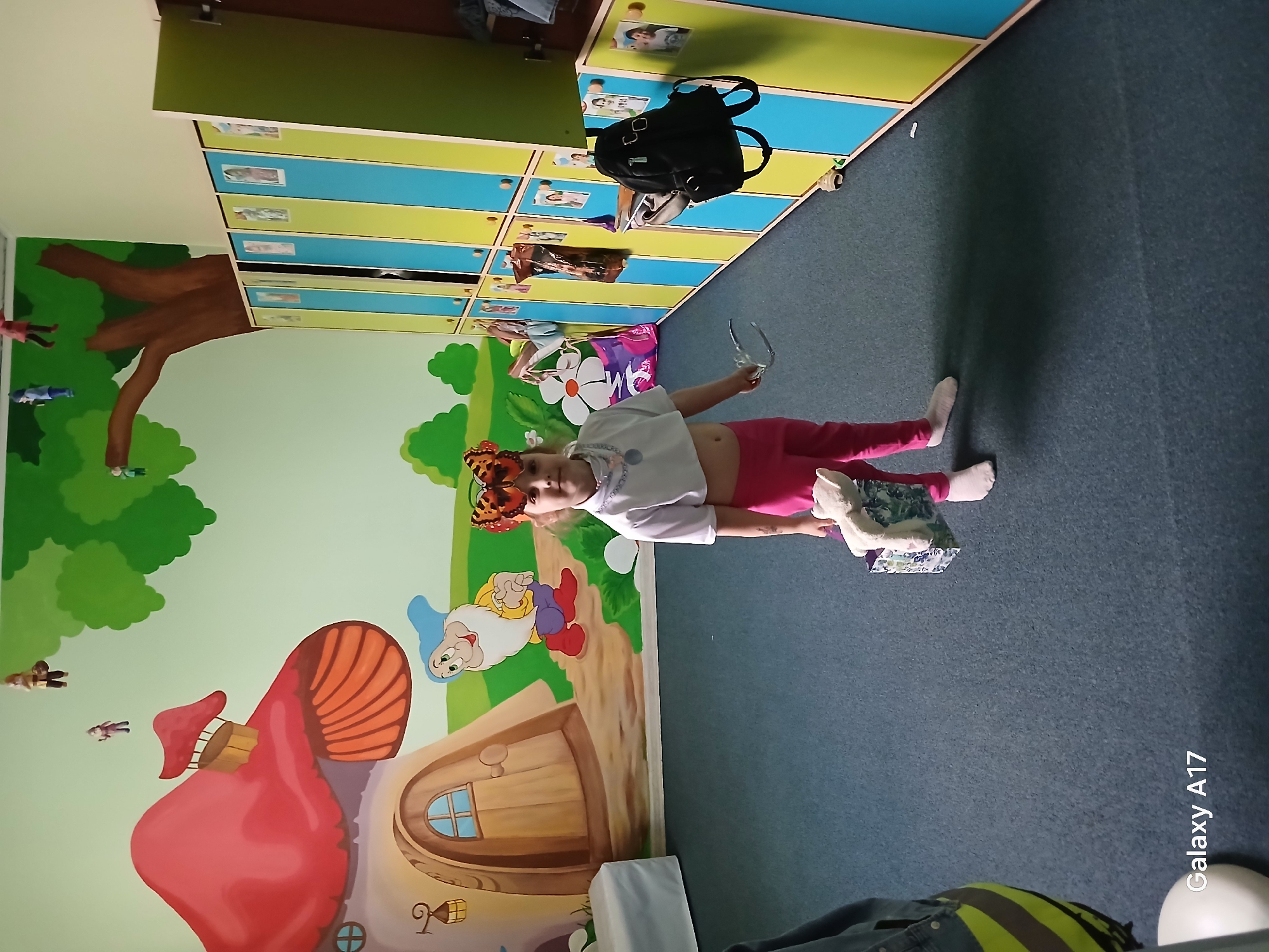 A young child with a butterfly mask stands in a room with a mural of a whimsical forest scene with mushrooms and a gnome, wearing a white shirt and pink leggings. The child holds a pair of glasses. Background includes lockers and a backpack. Location is potentially Yekaterinburg, Russia.
