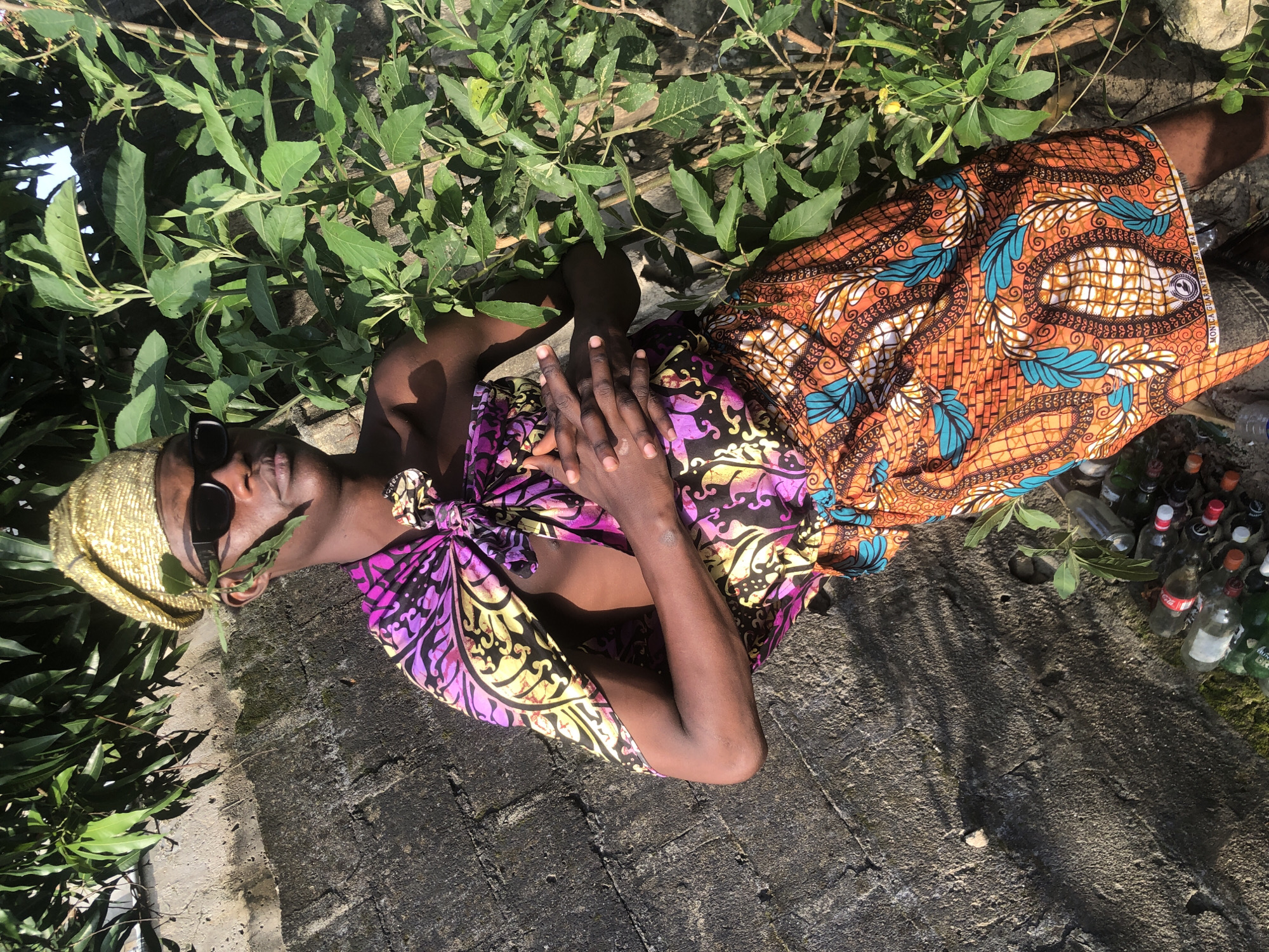 In Ado, Nigeria, a dark-skinned individual stands against a rough, grey, concrete wall, bathed in sunlight. The person wears a gold, textured headwrap and black sunglasses. A vibrant, patterned fabric wraps around their torso, featuring purple, yellow, and black designs, cinched with a knotted accessory. Below, another patterned fabric, predominantly orange with turquoise and white elements, is worn as a skirt. Green leaves from an adjacent bush are tucked into the headwrap, and more foliage grows to the right of the frame. In the lower left, a collection of plastic bottles lies on the ground. The scene is a close-up shot, focusing on the individual and their attire.