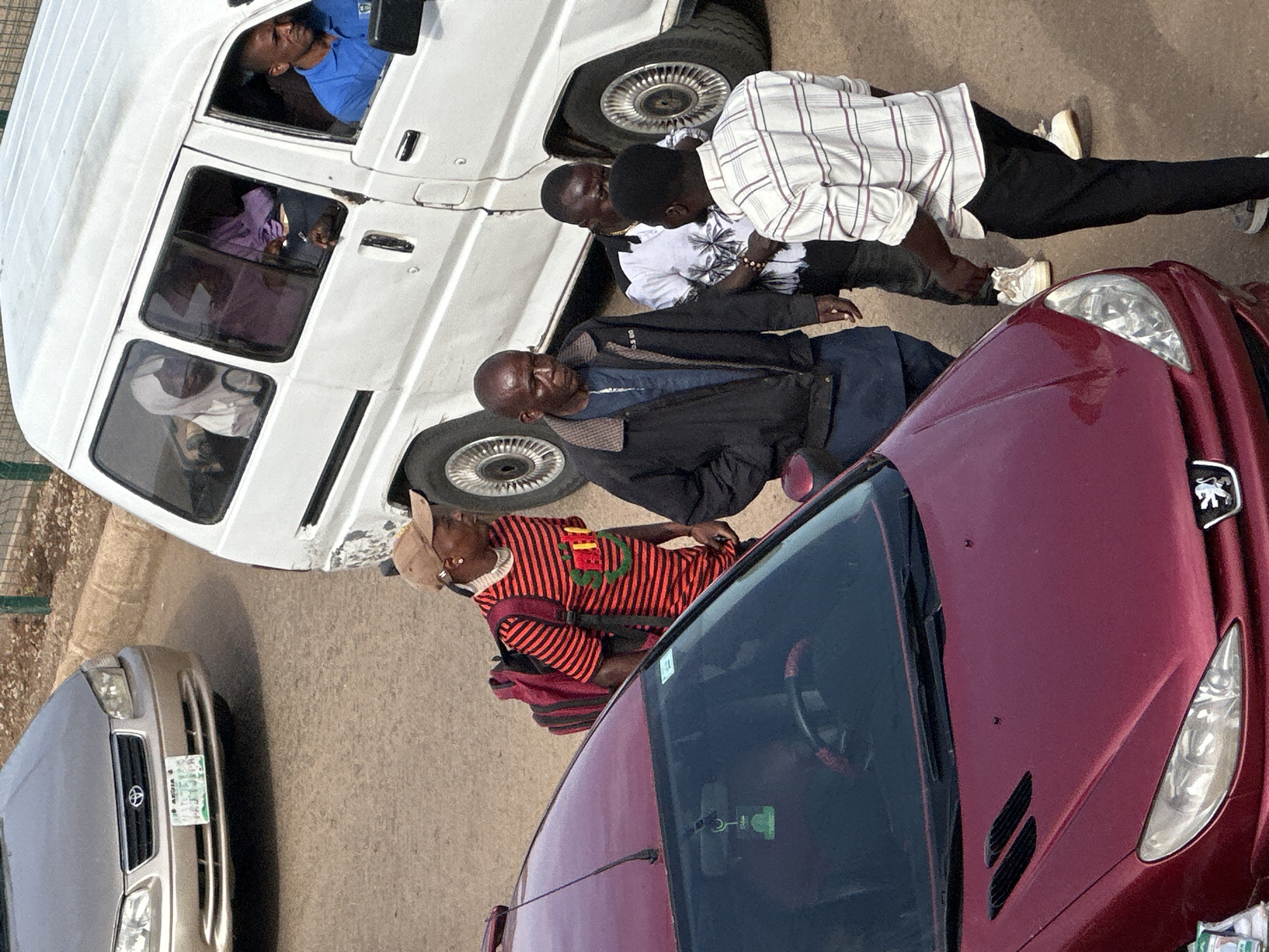 In Bukuru, Nigeria, on a paved road, a group of individuals are gathered near several vehicles. A burgundy-colored Peugeot sedan is in the foreground, positioned to the left. To the right of the sedan, a white van is parked, its passenger side facing the viewer. Inside the van, three individuals are visible: a driver wearing a blue shirt, and two passengers in the rear, one wearing a white headscarf and the other a purple garment.
Standing between the sedan and the white van are four men. The man closest to the sedan wears a red and black striped t-shirt and a tan baseball cap, carrying a red backpack. To his right, a man in a black jacket over a blue shirt is looking down. Next to him, a man in a white shirt with a palm tree pattern is interacting with another man wearing a white and purple striped shirt. The man in the striped shirt is facing away from the camera, and appears to be wearing black pants and white sneakers. Another car, a light-colored sedan, is partially visible in the upper left quadrant of the image. The ground is asphalt.