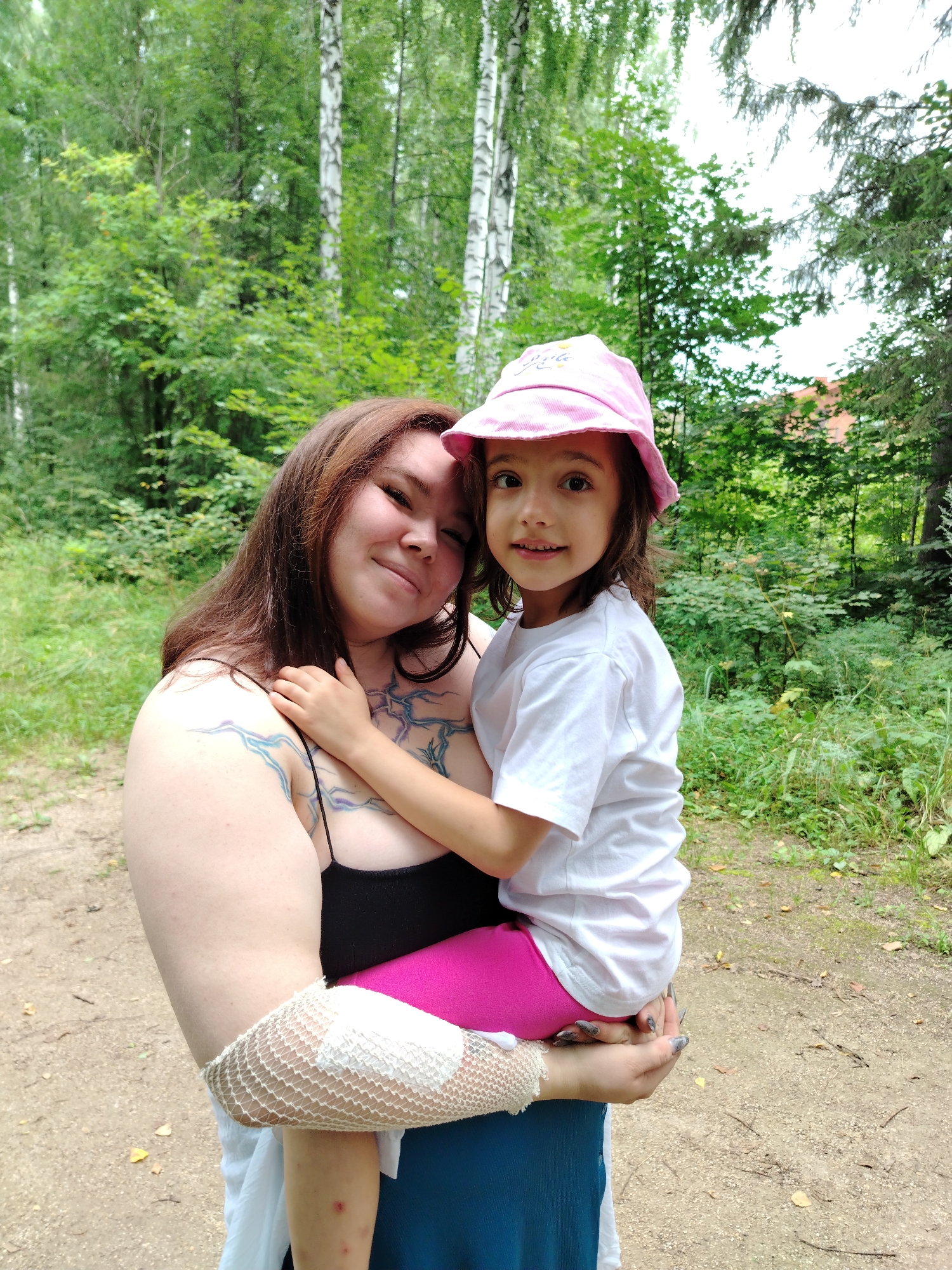 A woman is holding a young girl in her arms outdoors. The woman has long brown hair, tattoos on her arm and chest, and is wearing a black tank top and a blue skirt. She has a white mesh bandage wrapped around her left arm. The girl has dark hair, is wearing a pink bucket hat and a white t-shirt, and bright pink pants. They are in a wooded area with birch trees and green foliage visible. A dirt path is visible in the foreground. The location is identified as Noviy, Russia.