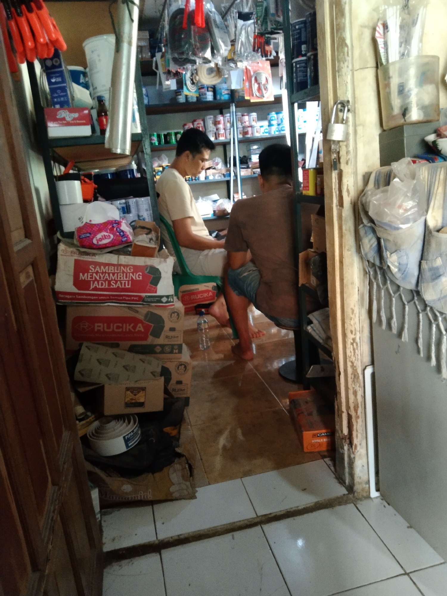 The image shows the interior of a shop in Parung, Indonesia, likely a hardware or plumbing supply store. The scene is cluttered with various items, including shelves stocked with goods, boxes stacked on the floor, and tools. Two men are present. One, wearing a light-colored t-shirt and white pants, is seated on a green plastic chair. The other man, wearing a brown t-shirt and denim shorts, is seated on a stool or a low platform, facing away from the camera. There are stacks of boxes labeled with "RUCKA" and other text, suggesting plumbing supplies. A roll of white tape is visible on the floor. In the background, shelves hold various containers and items, and a padlock is attached to a door frame. The overall impression is of a busy, functional retail space.