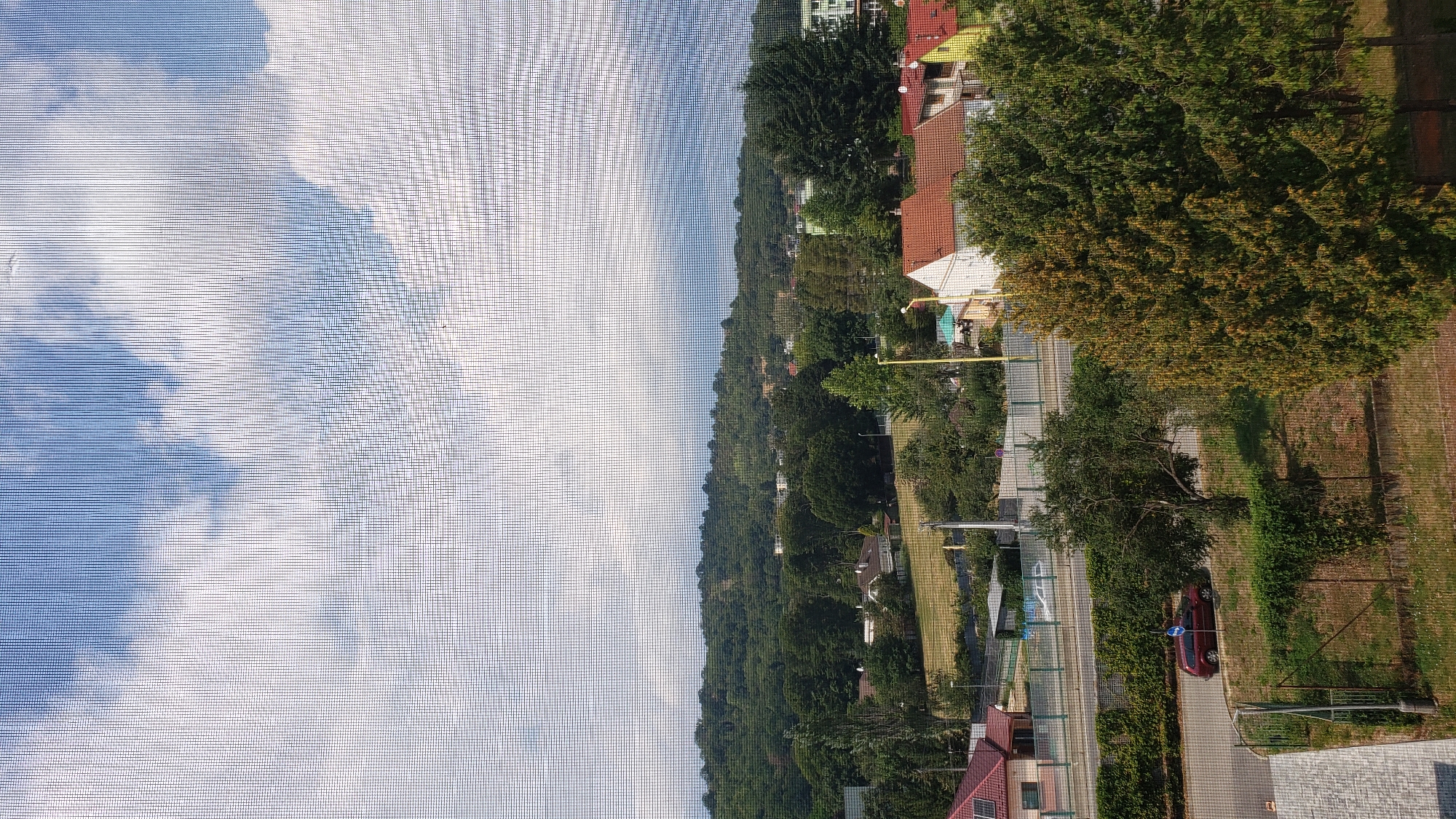 This image captures a view of a suburban area with residential houses, lush green trees, and rolling hills in the background, possibly taken from a window with a screen. The sky above is partly cloudy with a mix of blue and white. In the foreground, a red car is parked on the side of a road, with a pedestrian walkway and fencing visible. Several houses with red-tiled roofs are scattered throughout the landscape. The scene appears to be set in Prešov, Slovakia, a city known for its picturesque surroundings and a blend of urban and natural elements.