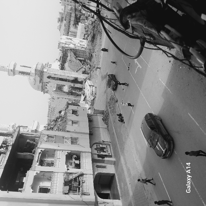 The image depicts a severely damaged urban street scene in Khan Yunis, Palestinian Territory, rendered in black and white. In the foreground to the left, a multi-story building, likely residential, exhibits extensive structural damage with large sections of its facade, windows, and balconies destroyed or missing, exposing interior rooms. Laundry hangs from a lower, intact balcony. A sign with a "Squid Game" triangle motif is visible on the ground floor. To the right, dominating the background, is a heavily damaged mosque with its minaret partially standing but leaning significantly. The mosque's dome and walls are extensively ruptured, and large piles of rubble surround its base. Other buildings in the background are also partially destroyed or reduced to debris.
A paved street runs horizontally across the mid-ground, featuring visible white lane markings. Two dark-colored cars are present on the street: one, a station wagon with white racing stripes on its hood, is in the foreground to the left, and another is further back near the mosque. Several individuals are dispersed throughout the scene. Three children sit on the curb to the left, near the damaged building. In the middle of the street, two figures, likely children, appear to be playing or interacting, one with arms raised. Other individuals, including children and an adult female, walk or stand further back on the street, near the mosque and the station wagon. Three figures walk on the left side of the street in the foreground, and one figure walks on the right side. The extreme right foreground shows out-of-focus wires and structural elements, suggesting the photo was taken from an elevated vantage point. The ground and sidewalks are littered with debris and rubble, consistent with heavy destruction. The sky is light and featureless.