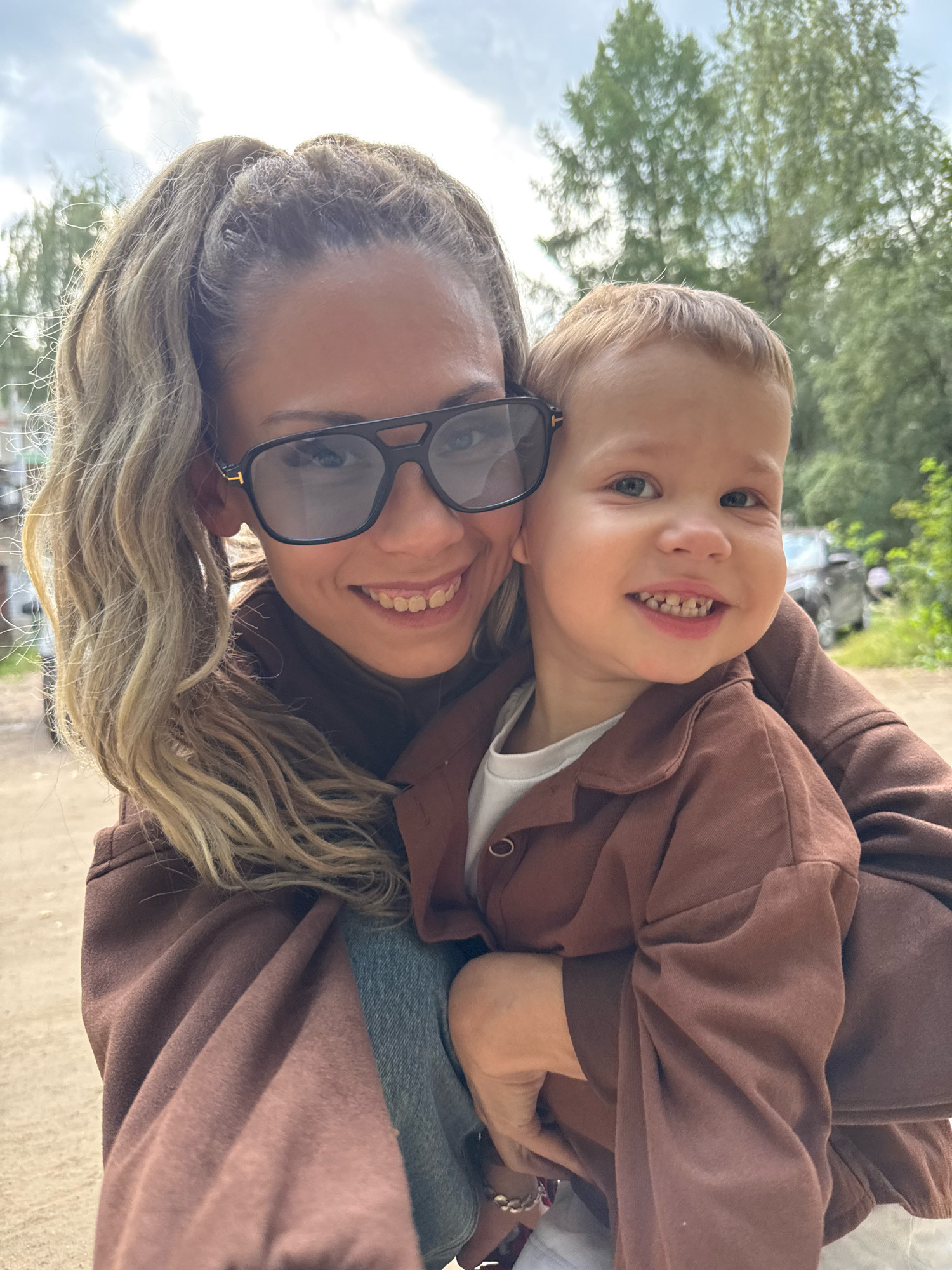 An outdoor, medium shot captures a woman and a young child in Rusanovo, Russia. The woman, with her blonde hair pulled back in a ponytail and wearing large, dark sunglasses, is smiling broadly, her teeth visible. She is dressed in a brown hooded sweatshirt. The child, who is positioned in front of her and being held, is also smiling and looking towards the camera. The child has light brown hair and is wearing a white t-shirt under a brown collared shirt. The background consists of trees and a cloudy sky, with a dirt path and a vehicle partially visible in the distance. The lighting suggests it is daytime.