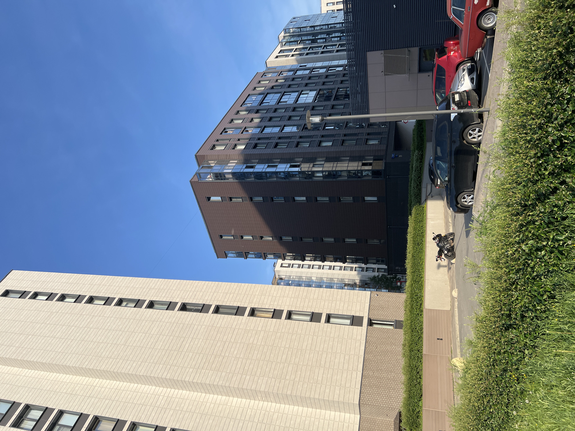 The image depicts a modern residential complex in Saint Petersburg, Russia, under a clear blue sky. In the foreground, lush green bushes and grass line a paved area. A parked motorcycle is visible on the pavement. Several cars are parked in a line: a black SUV, a silver sedan, a red SUV, and a vintage red car. Behind the parked vehicles, a dark, corrugated metal wall and a light brown building are present. In the middle ground, a dark brown apartment building with many windows and glass balconies stands tall, reflecting the sky. To its left, a lighter-colored, taller apartment building with numerous windows is also visible. Further in the distance, additional apartment buildings with white facades and glass elements can be seen. A street lamp stands near the brown apartment building. The overall scene suggests a well-maintained urban environment with contemporary architecture.