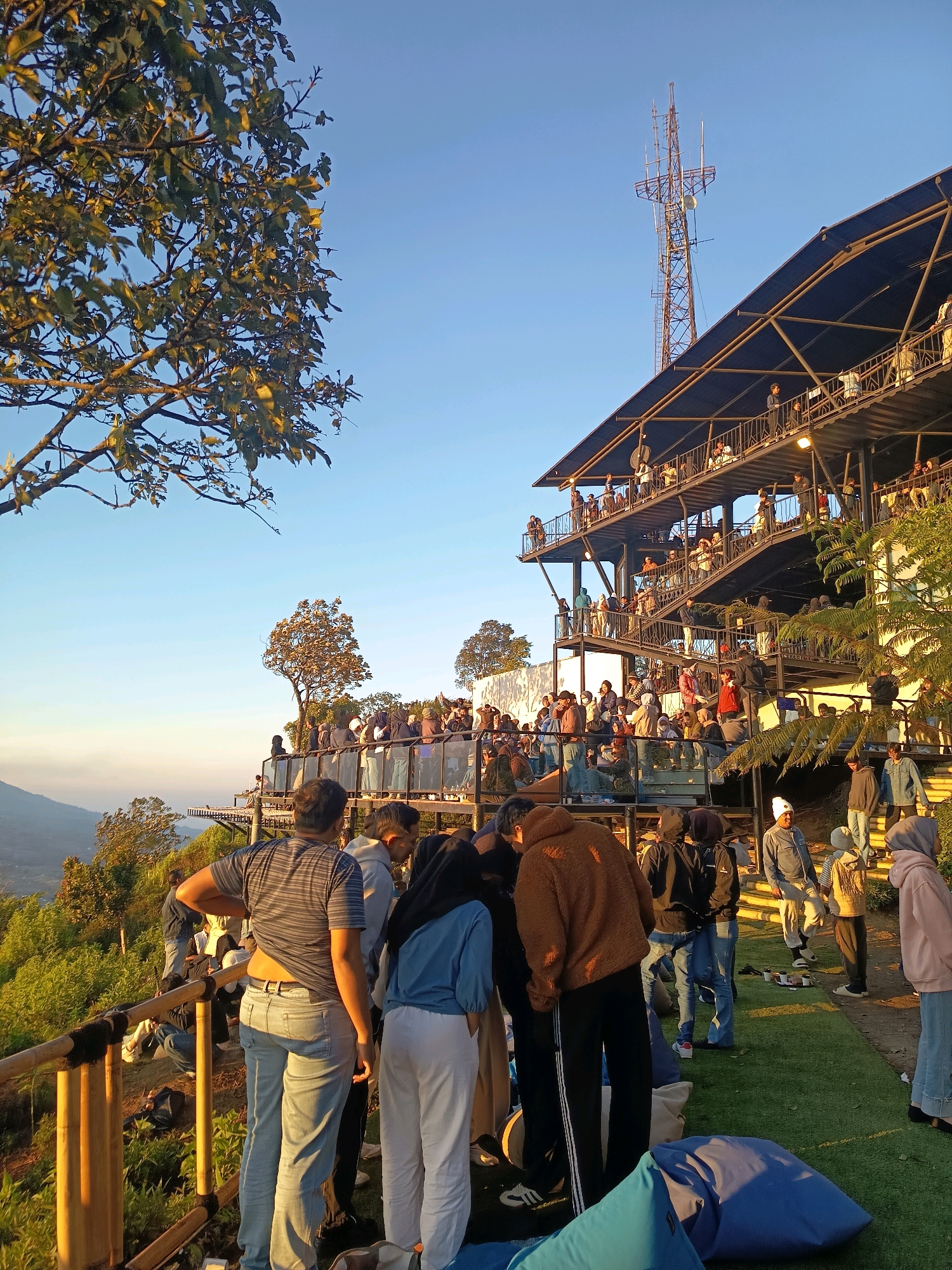 The image captures a crowded outdoor gathering on a multi-level structure in Keditan, Indonesia, during what appears to be late afternoon or early evening due to the warm lighting. The structure, resembling a tiered platform or building, is situated on a hillside overlooking a misty valley, with lush green vegetation surrounding it. Many people are visible on various levels, with some standing on balconies and walkways, while others are gathered on a grassy area in the foreground.
In the foreground, a group of individuals are clustered together. One person in a striped t-shirt and jeans is leaning against a bamboo railing, facing away from the camera. To their right, another person wearing a light blue top and white pants is also looking down. A person in a brown fuzzy hoodie is standing next to them, looking at something on the ground. Several beanbag chairs, one bright blue and another dark blue, are scattered on the green turf in the very front.
The tiered structure behind the foreground group features multiple levels with railings and open walkways, supporting a significant number of people. People are seen walking on stairs and standing along the edges, observing the surroundings. A tall communication tower stands prominently against the clear blue sky behind the structure, adding a distinctive industrial element to the natural landscape. The overall scene suggests a popular viewpoint or recreational area, possibly a cafe or restaurant with observation decks, experiencing high foot traffic. The ambiance is lively, with a mix of people dressed in casual attire, indicative of a relaxed social event.