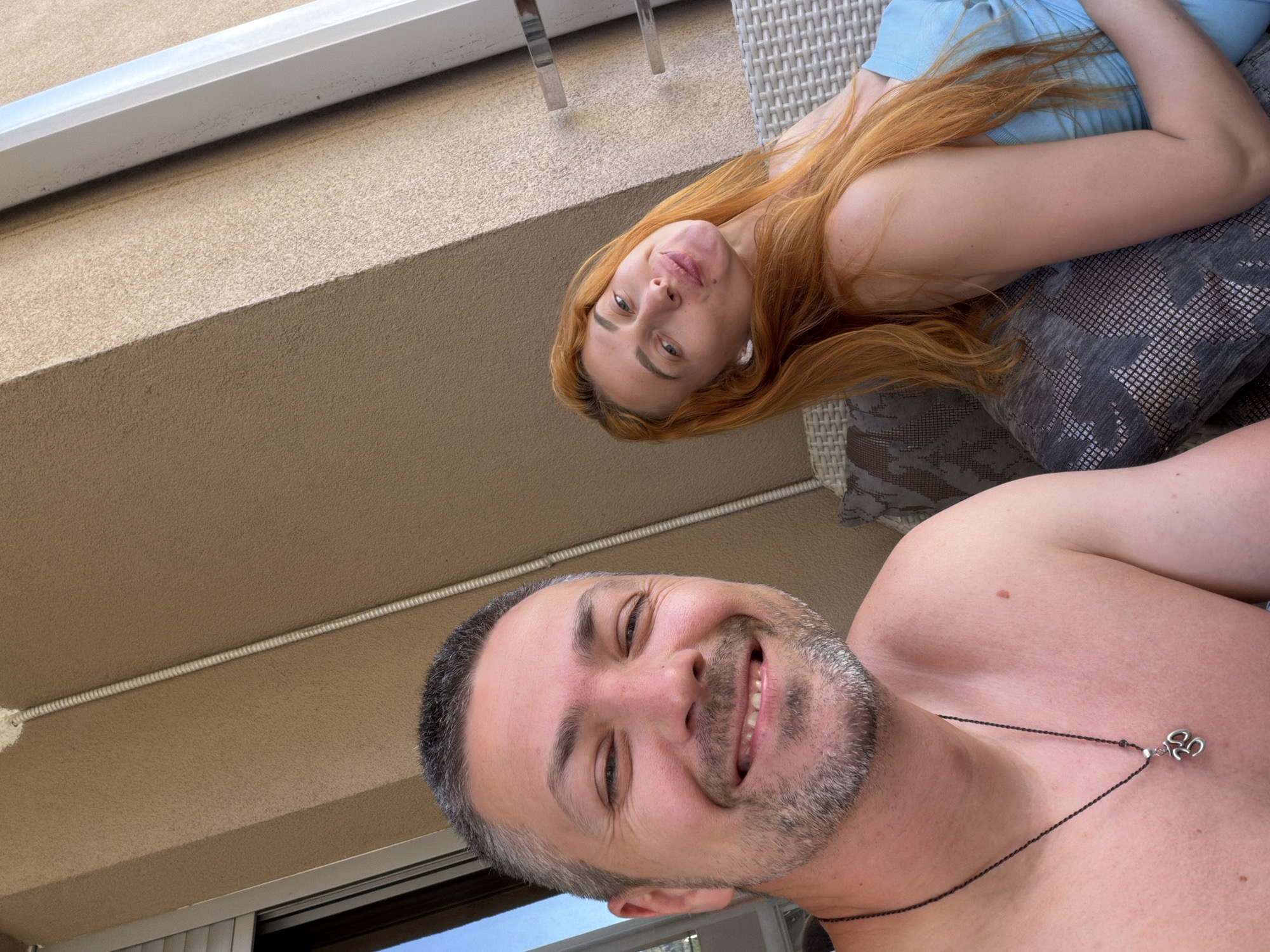 The media file is a selfie featuring a man and a woman. The man is in the foreground, looking directly at the camera with a wide smile. He has short, graying hair and a goatee. He is shirtless and wearing a dark necklace with a clover-shaped pendant. The woman is lying down behind him, to the left side of the frame, looking upwards with a slight smile. She has long, reddish-orange hair and is wearing a light blue top. The background appears to be an outdoor setting, possibly a balcony or patio, with a beige textured wall and a white railing visible. The furniture suggests a relaxed setting, likely a lounge area. The lighting indicates it is daytime, with natural light illuminating the scene. The overall mood is casual and happy.