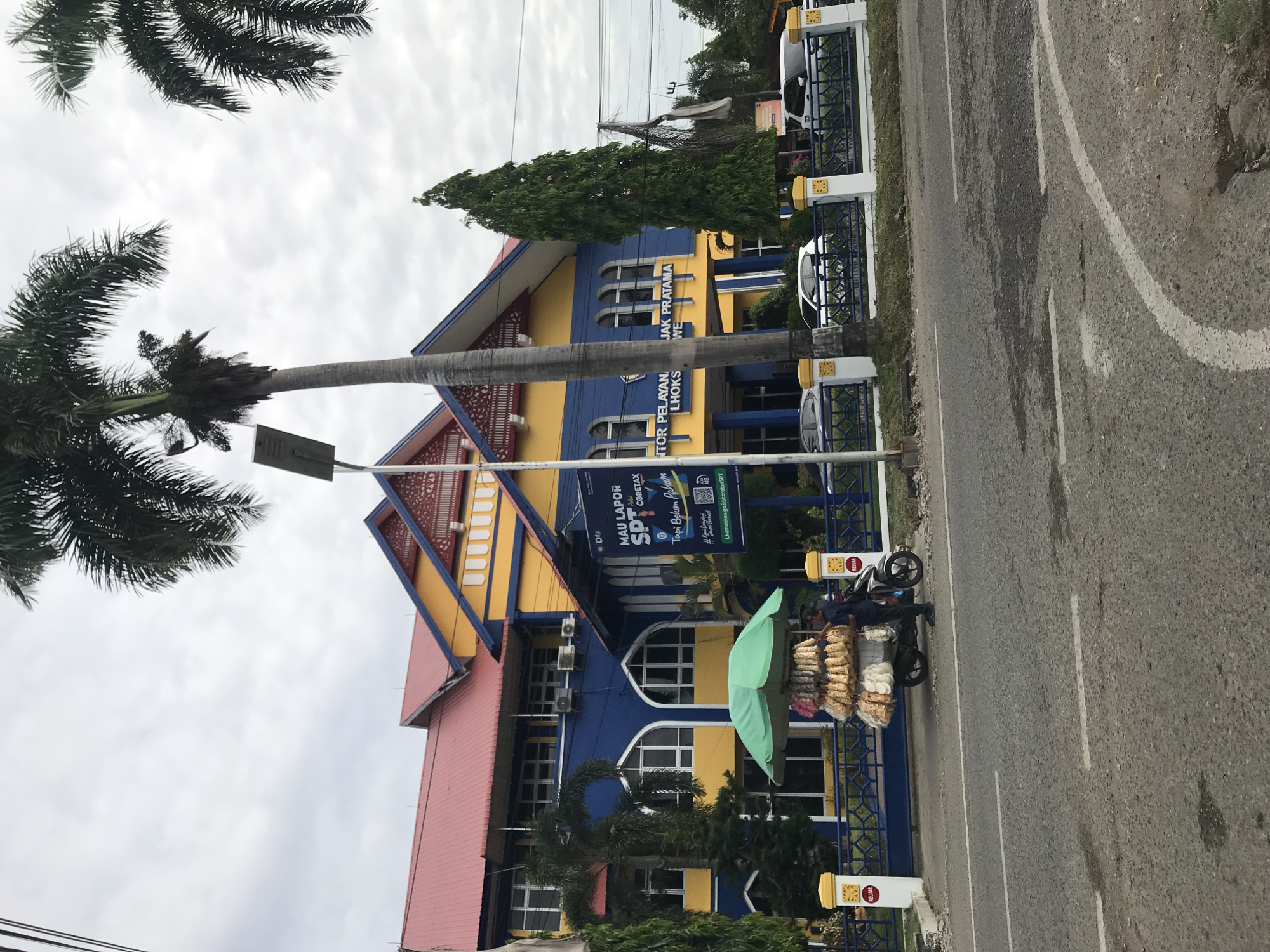 The image depicts the exterior of the Kantor Pelayanan Pajak (KPP) Pratama Lhokseumawe (Tax Service Office) in the city of Lhokseumawe, Indonesia. The building is predominantly blue and yellow, with a red roof, and is adjacent to a street. A palm tree is visible in the foreground. A small vendor is set up near the building selling snacks or food items from a motorcycle. Several cars are parked near the building. Banners and signage related to taxation and reporting (SPT CoreTax) are displayed. The sky is overcast.