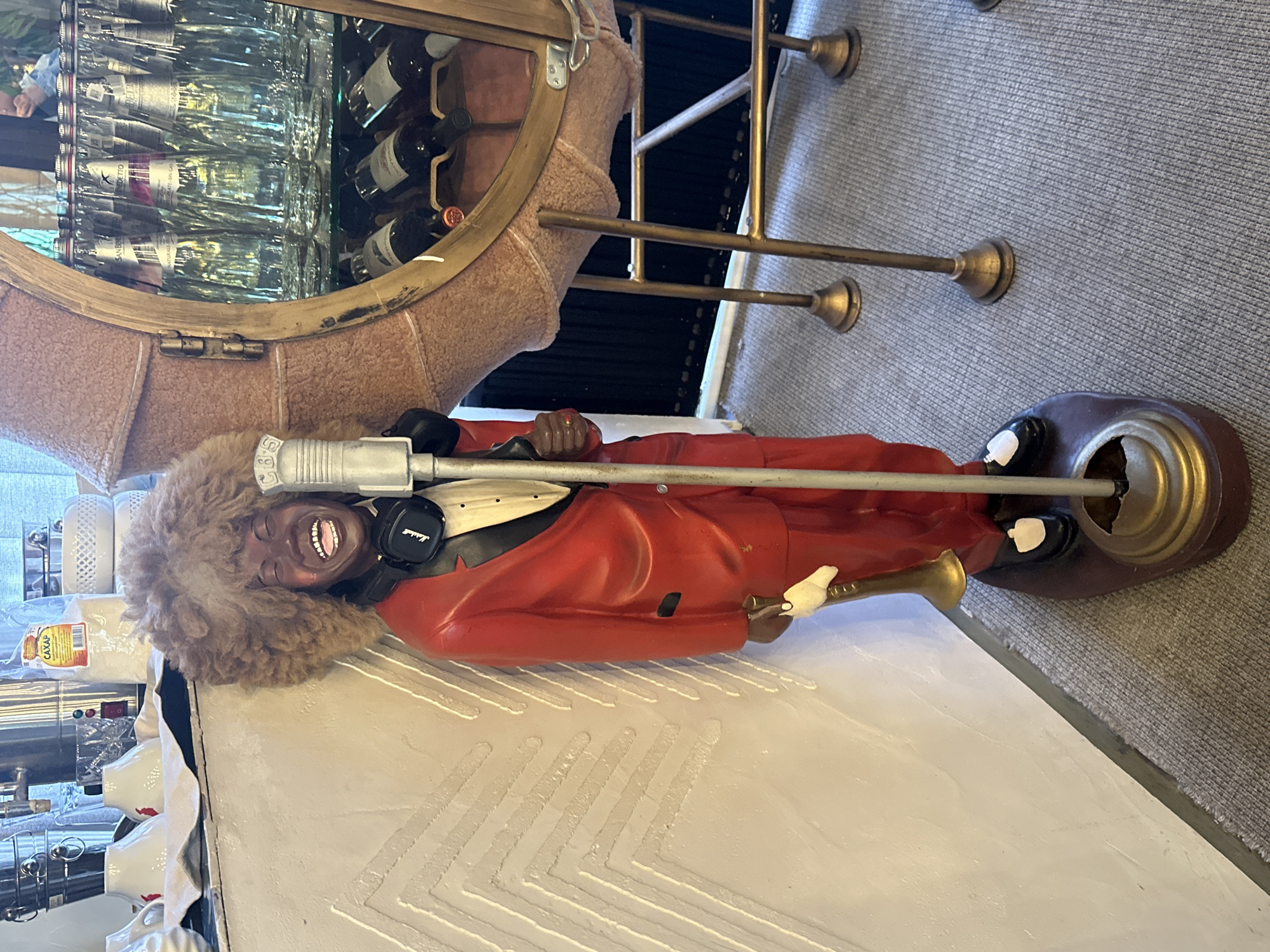 The image shows a life-size statue of a Black man wearing a red suit, a bow tie, and headphones, holding a trumpet and standing next to a microphone. The statue has a large, frizzy brown afro and is laughing. Behind the statue, a round, gold-trimmed cabinet with mirrored doors displays numerous bottles of wine and other beverages. The cabinet is supported by four gold-colored legs. The statue stands on a dark, oval base, which is placed on a grey carpeted floor. To the left of the statue, a white wall features a textured, zigzag pattern. In the background, to the left of the statue's head, several white ceramic cups and a bag of sugar are visible on a counter. The environment appears to be an indoor establishment, possibly a restaurant or bar. No people are present in the scene. The location of посёлок гидроузла имени Куйбышева, Russia, is not discernible from the visual information provided in the image.