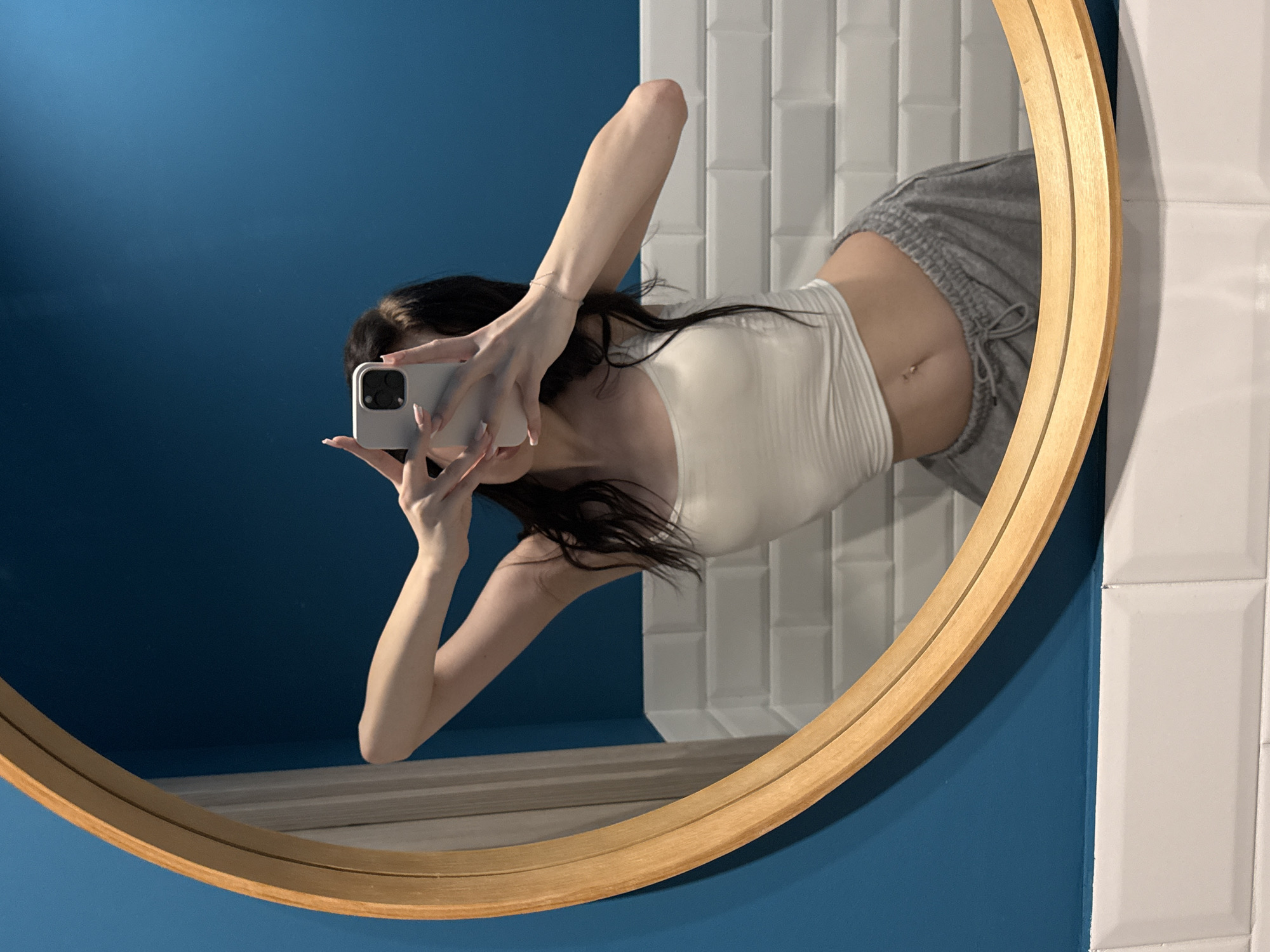 The image shows a young woman in a white crop top and grey sweatpants taking a mirror selfie. She has long dark hair and her belly button piercing is visible. The background consists of a blue wall and white tiled sections. The mirror is round with a wooden frame. The image is taken indoors, likely in a bathroom. The location of Zakharovo, Russia is not depicted in this image.