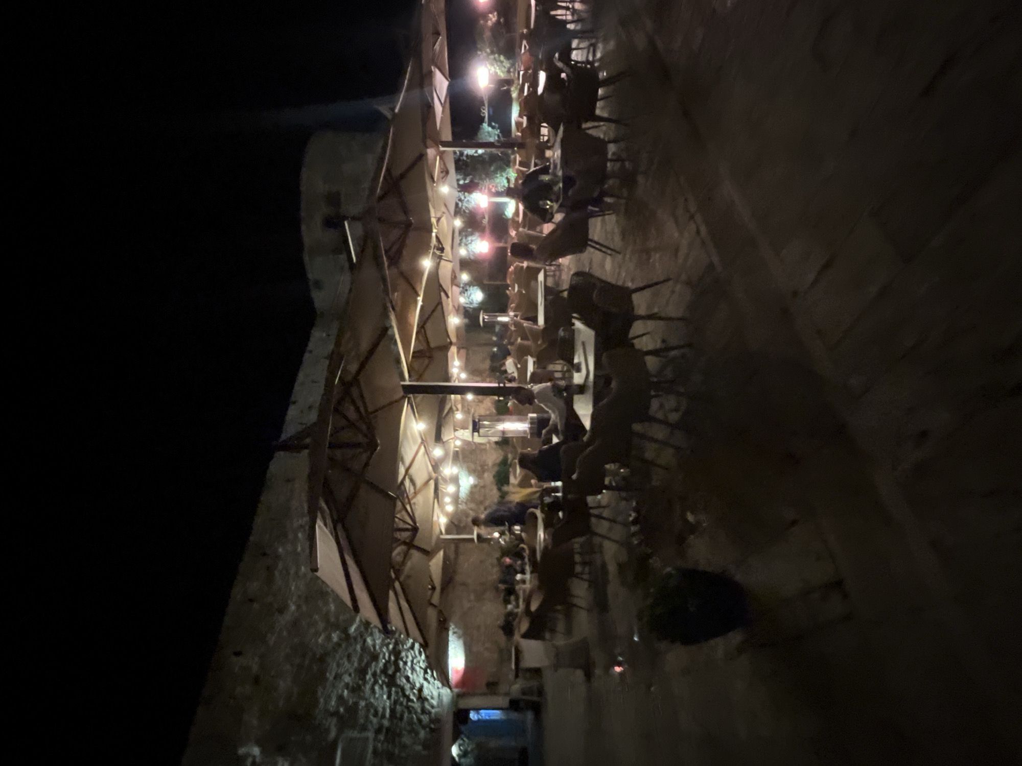 The image depicts an outdoor dining area at night, possibly in Budva, Montenegro, given the stone architecture. The scene is dimly lit with string lights and spotlights illuminating the tables and surrounding structures. Numerous people are seated at tables, suggesting a restaurant or café setting. The tables are covered, and chairs are arranged around them. The area is covered by a canopy, likely providing shelter from the elements. A stone wall is visible on the left side, adding to the ambiance. A large, dark, indistinct object sits on the right side.