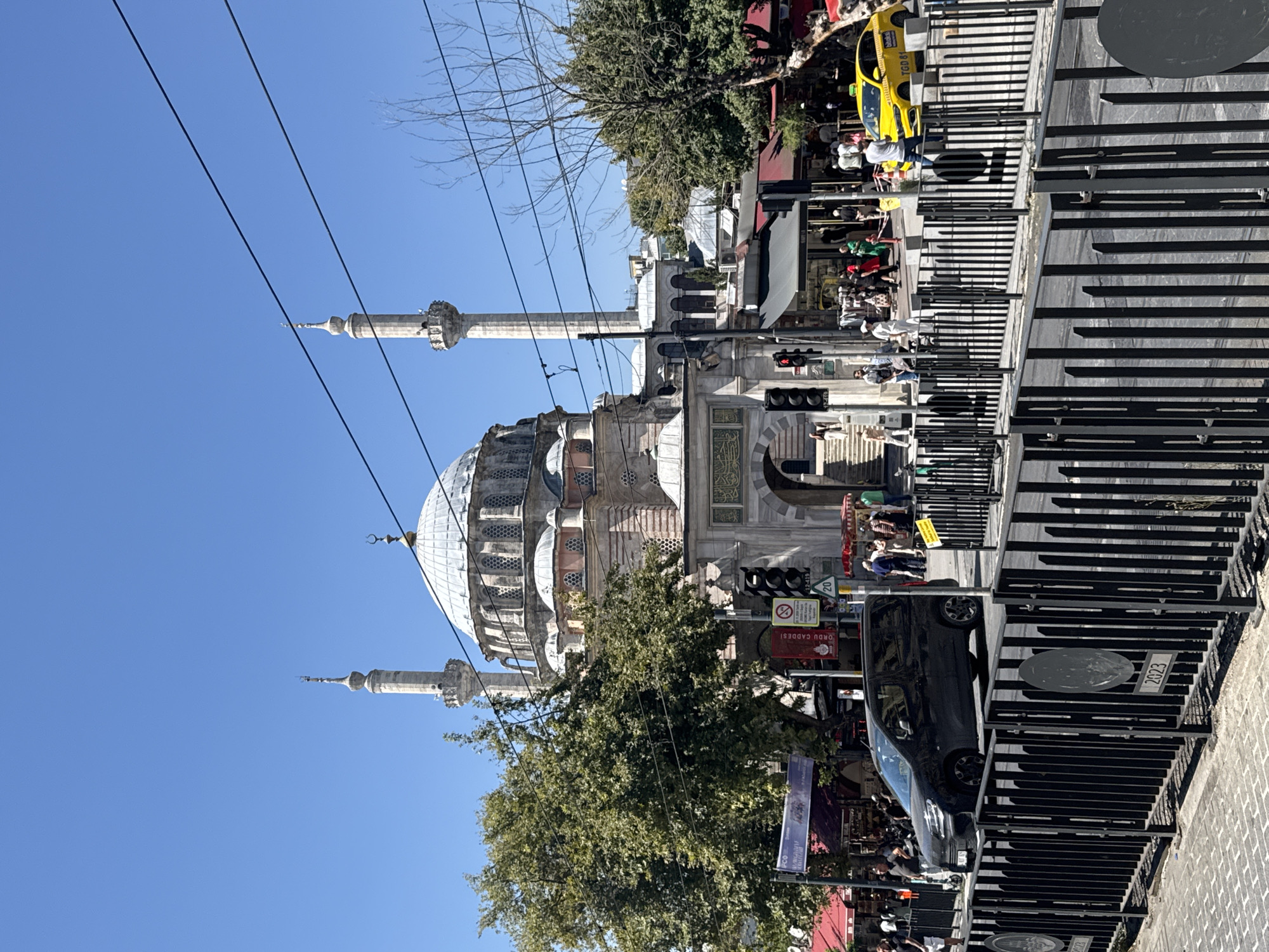 The image captures a view of the Ortaköy Mosque in Istanbul, Turkey, on a bright, sunny day. The mosque, a prominent landmark, features a large dome and two minarets. In the foreground, a black minivan is parked on the street. A yellow taxi is also visible, further down the road. People are scattered around, some walking on the sidewalk, others appearing to be patrons of the shops lining the street. Red awnings of shops are visible, suggesting a bustling commercial area. A black metal fence separates the street from a paved walkway or pedestrian area. Traffic lights are visible, indicating an intersection. The sky is clear blue.