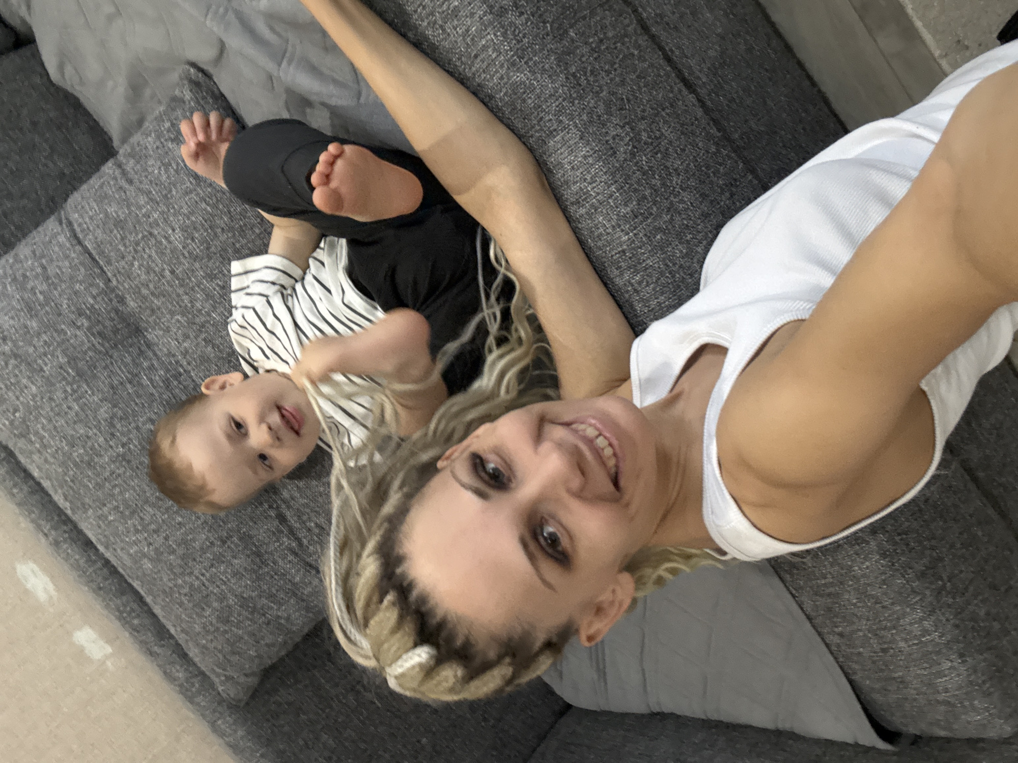 A woman and a young child are lying on a gray sofa. The woman, wearing a white tank top, has blonde hair styled in braids. She is smiling and looking directly at the camera, holding it with her right hand. The child, wearing a striped shirt and dark pants, is lying next to her, with their bare feet visible. The sofa appears to be in a home setting. There is no evidence to support that this is in the city of Rusanovo, Russia.
