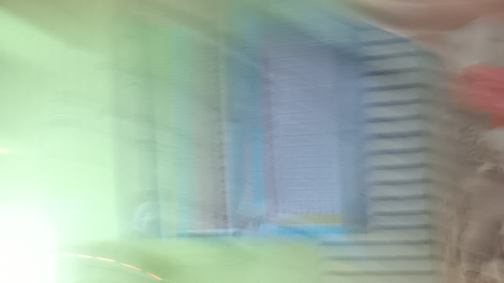 The image, taken in Sonobekel, Indonesia, is a blurry, abstract composition dominated by soft, pastel colors and diffused light. Vertical elements, suggestive of a fence or railing, are visible in the lower portion of the frame, colored in muted grays. Above this, horizontal bands of color are present: a light blue band is prominent, with a thin gray band above it and a more vibrant blue band beneath. The upper half of the image is a wash of pale green, transitioning to a bright, almost white, upper edge. The overall impression is one of movement and a lack of sharp focus, making specific identification of subjects or objects difficult. There is no discernible interaction or activity captured due to the blurriness.