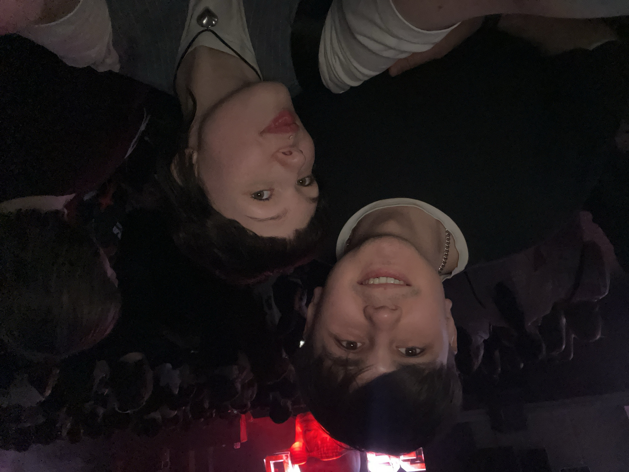 Two individuals are visible in the foreground of the image, with a crowd of people in the background. The lighting is dim, with a red light source illuminating the lower portion of the scene. The location appears to be a concert or event. The image is oriented upside down, making it difficult to discern further details about the location in Kazan, Russia.