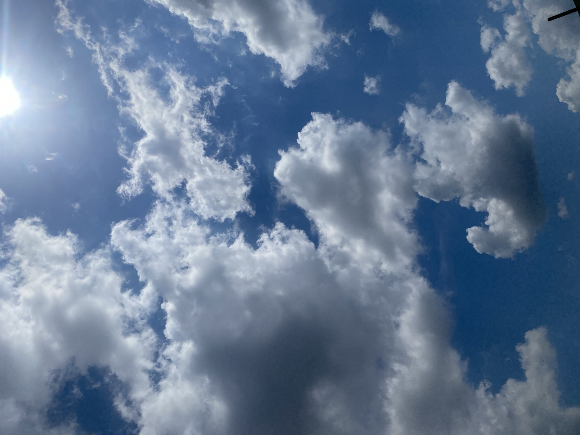 An extreme upward-facing shot captures a brilliant blue sky dotted with fluffy, white cumulus clouds. Sunlight streams in from the top right, creating a slight lens flare and casting a soft glow on the clouds. The clouds are varied in size and density, with some appearing dense and shadowed, hinting at their volume. The image is taken from Berangas Timur, Indonesia.