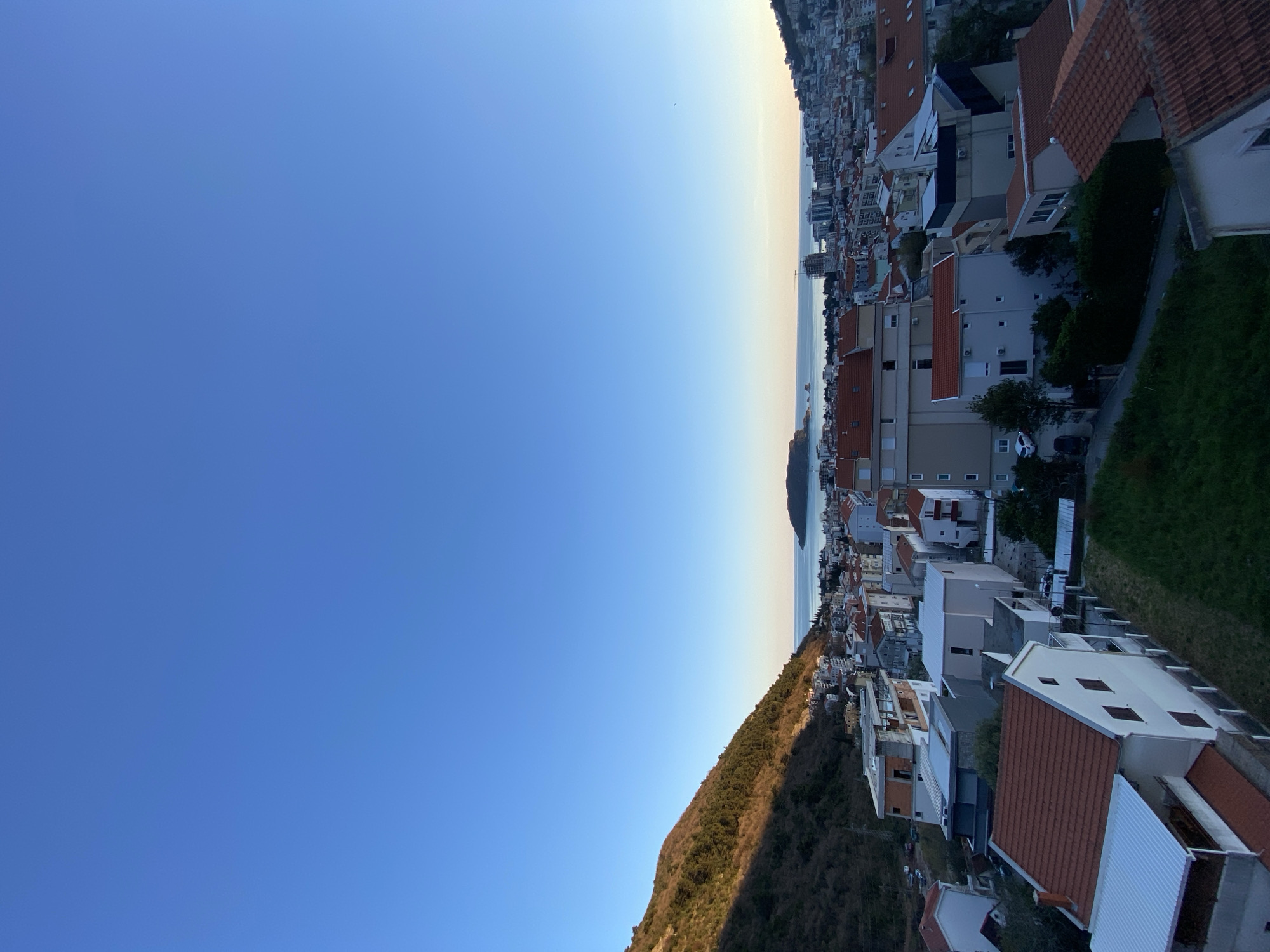 The image shows a partial view of the city of Budva, Montenegro. The foreground is a blue sky that fades to white at the horizon. A hill covered with sparse vegetation is visible. Buildings with red and white roofs are clustered together, some appearing to be residential, and others are taller and more modern. A body of water, likely the Adriatic Sea, is visible between the buildings. An island is visible at a distance in the water.