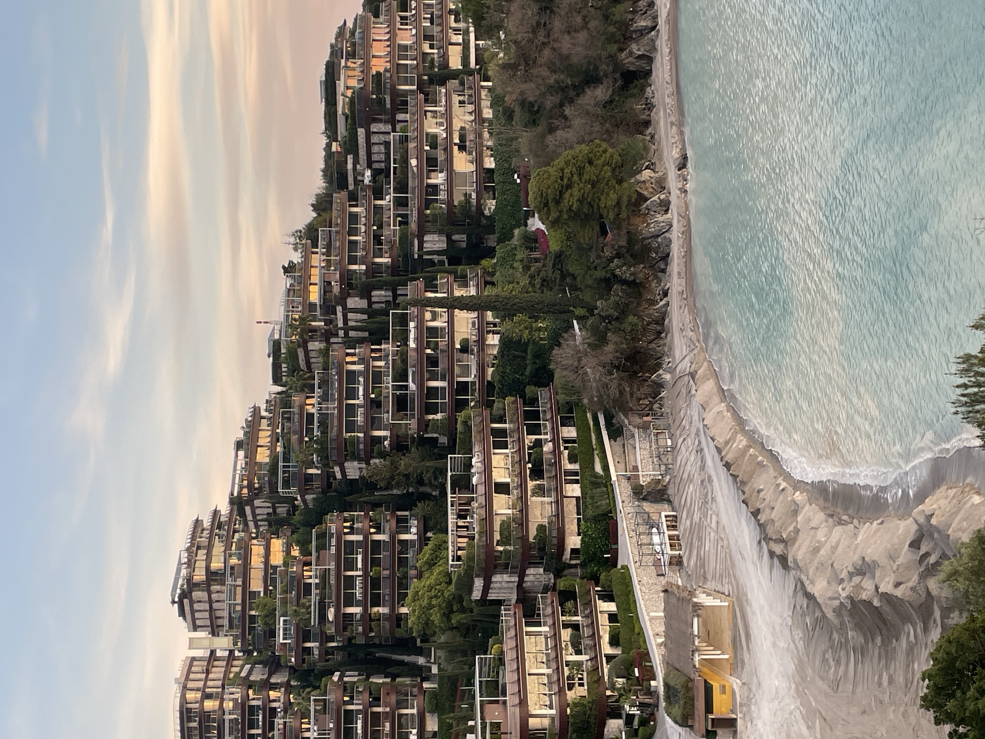 An elevated, wide-angle view depicts a coastal scene in Budva, Montenegro, specifically identifying the multi-tiered Dukley Gardens complex on the Zavala Peninsula. The scene is illuminated by soft, warm light, indicating either dawn or dusk.
The foreground features a crescent-shaped sandy beach fronting a calm bay with clear, light turquoise water. The sand on the beach is arranged in distinct, undulating rows parallel to the waterline. At the left end of the beach, a small yellow building with a thatched roof is visible. Further right, a rectangular, open-frame structure stands on the sand, containing a white board, likely a surfboard or paddleboard. A stone retaining wall separates the beach from a paved path that ascends the hillside.
The midground and background are dominated by numerous multi-story residential buildings, terraced into the steep, green hillside. These modern structures feature light-colored facades, dark brown or bronze accents, extensive glass windows reflecting the warm sky, and numerous balconies with visible outdoor furniture. Many rooftops are flat and incorporate greenery or additional terraces. Lush green vegetation, including tall, slender trees (likely cypresses), and various shrubs, densely surrounds and intersperses the buildings.
The sky above is light blue, transitioning to soft pink and orange hues near the horizon, with sparse, wispy clouds. No individuals are visible anywhere in the scene.