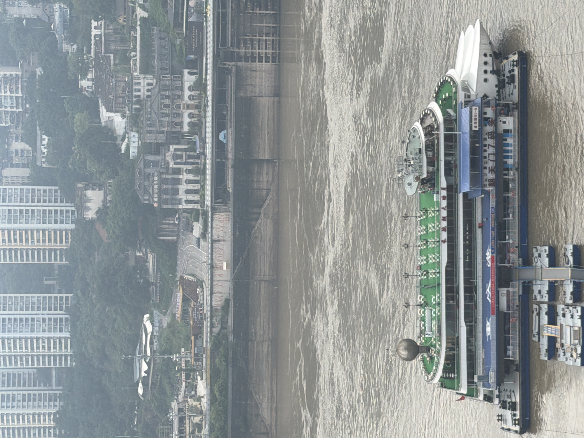 In Yuzhong District, China, a large, multi-level cruise ship is docked in a wide, muddy river. The ship is white with green decks and blue lower levels, featuring numerous windows and railings. Several smaller barges are attached to the side of the cruise ship, connected by gangways. In the background, a concrete embankment lines the river, with a road and buildings situated on a hill above. The cityscape is characterized by dense clusters of modern high-rise apartment buildings, interspersed with older, more ornate architecture. Dense green foliage covers the hillsides, partially obscuring the buildings. The sky is overcast, contributing to a hazy, muted atmosphere. No people are clearly visible in the scene.