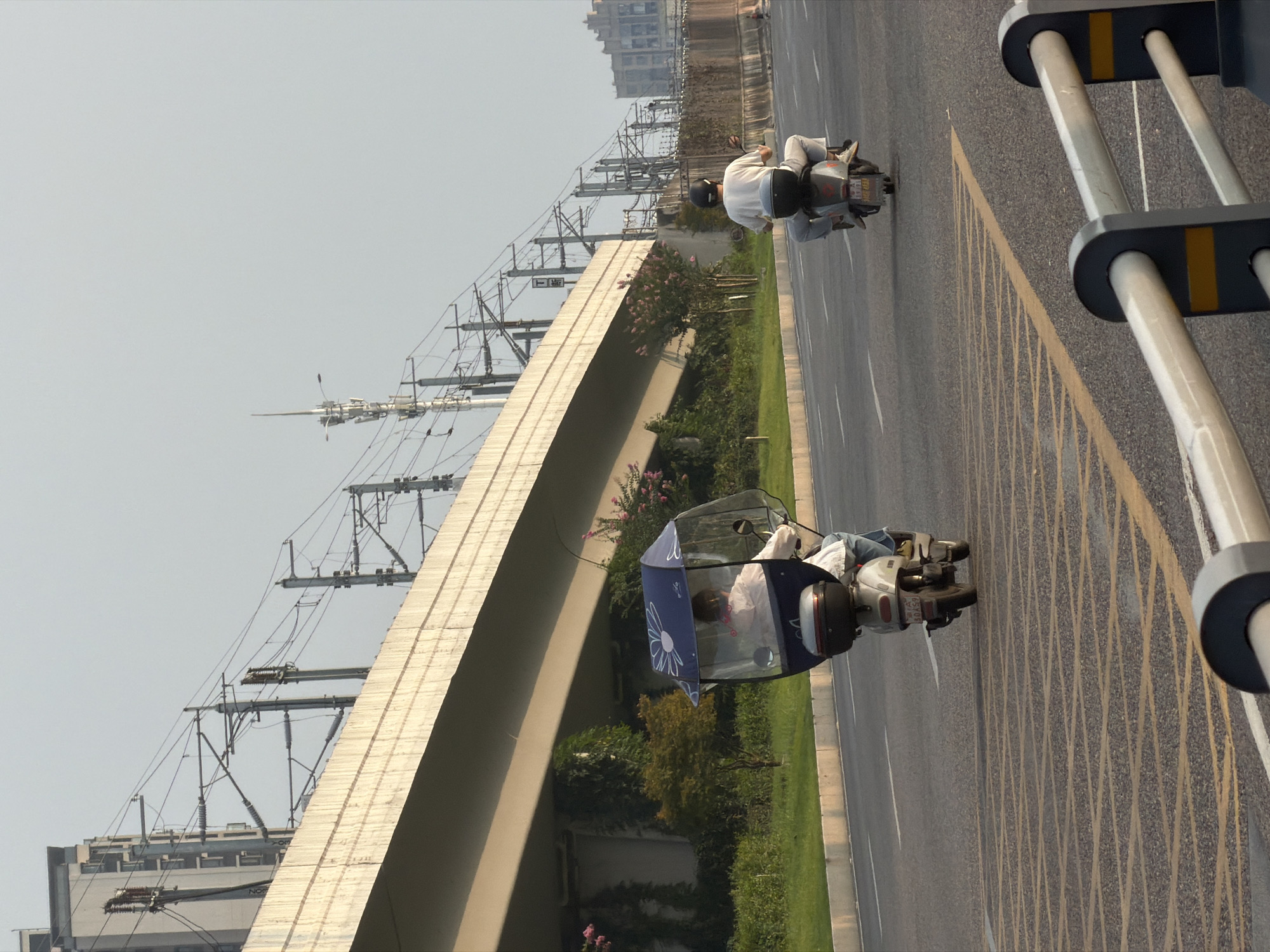 The image shows two people riding motor scooters on a paved road in Jiangjin, China. The road has lane markings and a yellow crosshatch pattern in the foreground. In the background, there is an elevated highway or bridge structure with overhead power lines and poles. A building is visible behind the power lines to the left, and another building is visible in the distance to the right.
The scooter on the left is being ridden by a person who is covered by a transparent blue canopy with a white flower pattern. This rider is wearing a white top and jeans.
The scooter on the right is being ridden by a person wearing a white t-shirt and light-colored pants, with a black helmet. This scooter has a black storage box on the back. Both scooters have license plates.
The environment appears to be a roadside with green grass and some trees and bushes along the embankment of the elevated structure. The sky is overcast.
