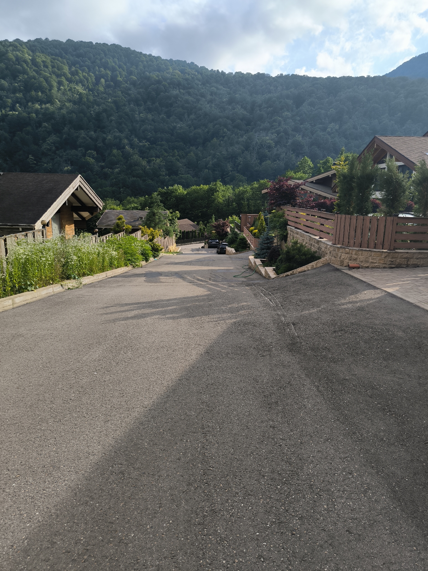 A paved road winds uphill through a residential area in Krasnaya Polyana, Russia, with dense, green forest covering the steep mountainside in the background. The sky above is partly cloudy. On the left side of the road, a wooden fence separates the asphalt from a row of houses with dark roofs and white-flowered bushes. On the right, a stone retaining wall topped with a wooden fence borders the road. A black car is parked further up the road. The scene appears to be a quiet, upscale neighborhood nestled in the natural beauty of the mountains.