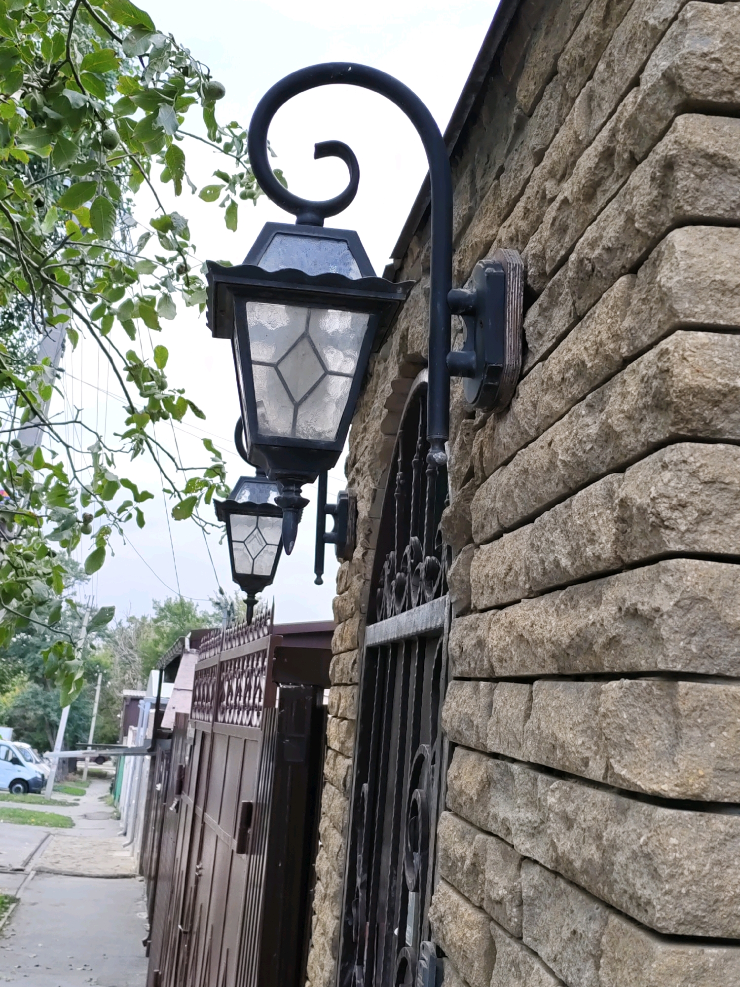 The image depicts an outdoor scene in Rostov-on-Don, Russia. In the foreground, a textured stone wall is visible, constructed with horizontally laid rectangular stones. Mounted on the wall are two ornate black metal lanterns, one above the other, with glass panes. A decorative, curled metal arm supports the upper lantern. To the left of the wall, a wrought iron gate is partially visible. Beyond the gate, a paved walkway runs alongside a row of dark brown metal gates and fences. Trees with green leaves are present on the left side of the image, with some branches extending over the wall and lanterns. In the background, a street with parked vehicles, including a white van, and further fences and buildings are discernible under an overcast sky. The scene appears to be a residential or commercial area.