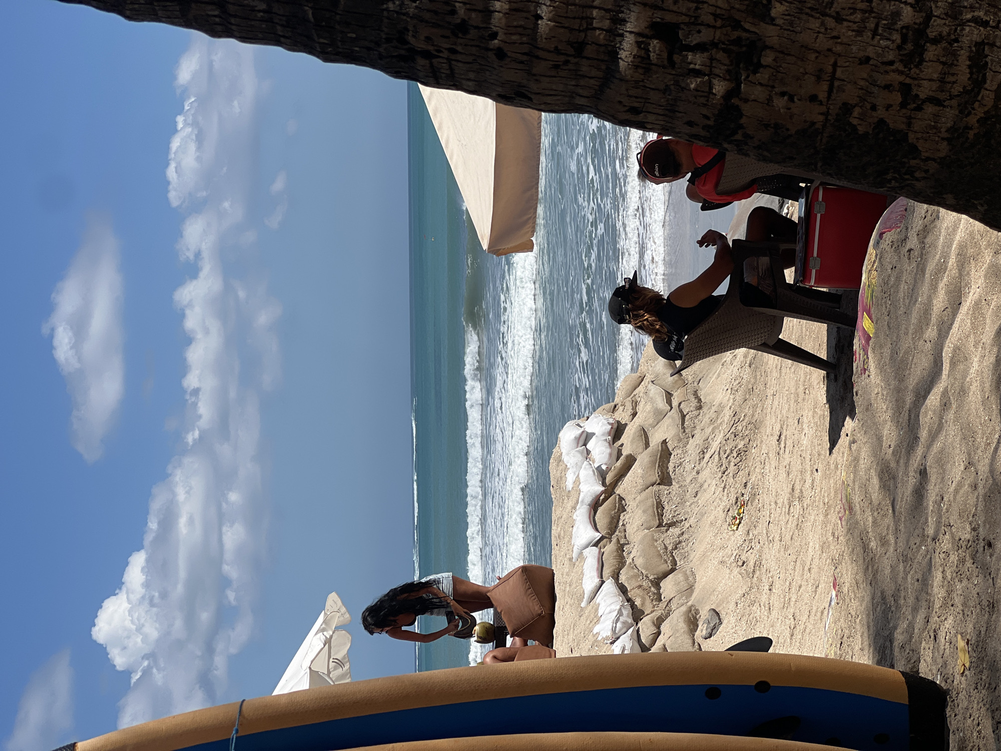 This image depicts a beach scene in Kuta, Indonesia, on a bright, sunny day with a clear blue sky and scattered white clouds. The ocean in the background has moderate waves. In the foreground, a large surf board is leaning against a sandbagged embankment.
To the left, a woman with long dark hair stands near a white umbrella, holding a coconut drink. She is wearing denim shorts and a bikini top. A brown beanbag chair is situated behind her.
In the middle ground, two people are seated in chairs facing the ocean. The person on the left, wearing a black baseball cap and a black tank top, is holding something in their hand. A red cooler box is placed on the sand in front of them. The person on the right is wearing a red life jacket or vest and a black cap.
The sandbags forming a barrier on the embankment are a prominent feature. The area behind the people seated in chairs appears to be a sandy beach with some debris. A palm tree trunk is visible on the far right, framing the scene.