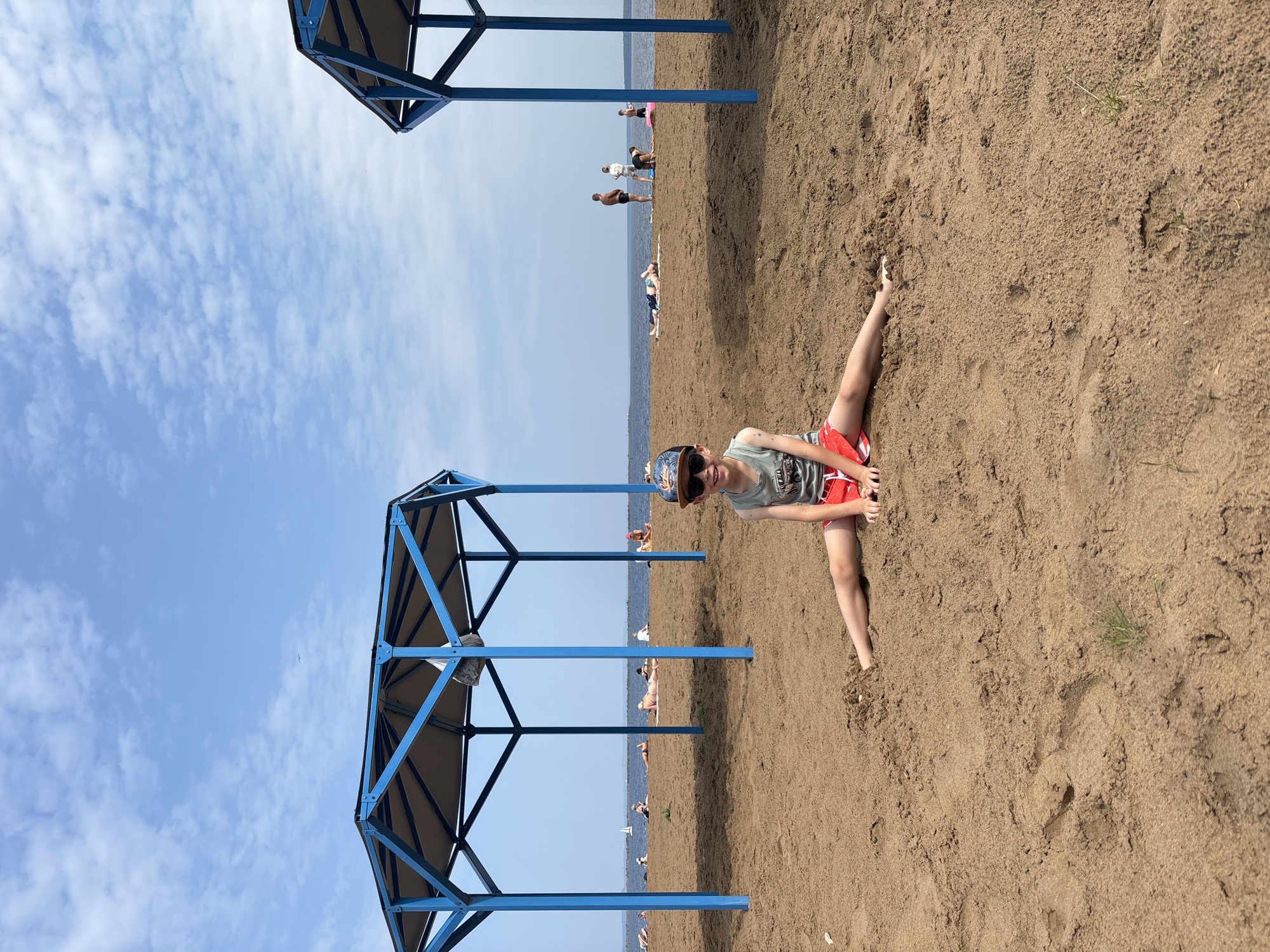 A young boy with sunglasses and a baseball cap sits on a sandy beach in Chaikovsky, Russia, in a split position. He is wearing a grey tank top and red shorts. Two blue canopies provide shade on the beach. Several people are sunbathing and walking in the background near the water. The sky is blue with scattered clouds.