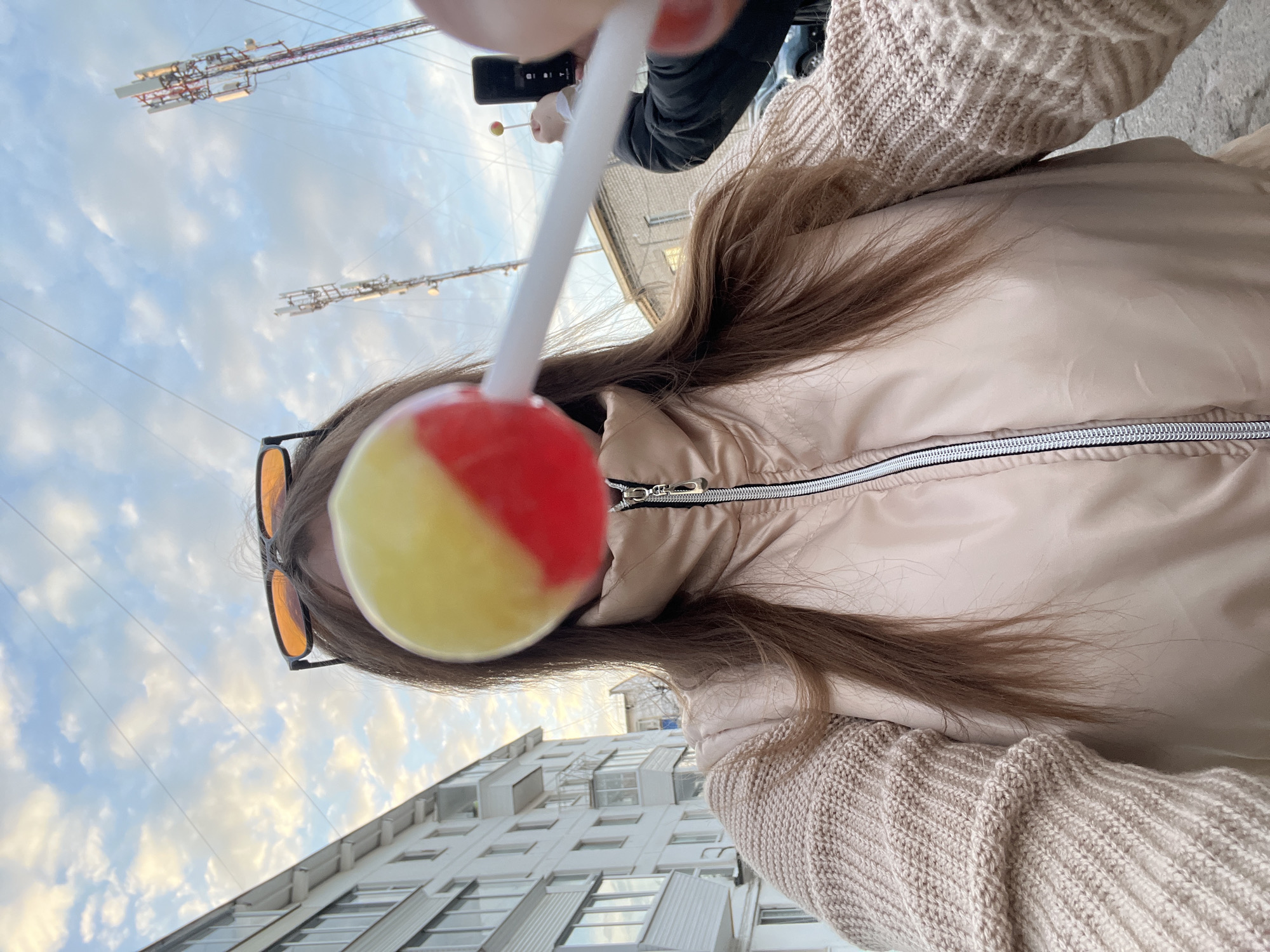 A selfie-style, low-angle shot shows a person holding a lollipop in front of their face, obscuring the features. The lollipop is round and divided into yellow and red halves. The person wears orange-tinted sunglasses, a beige puffer jacket, and a beige knitted sweater. Long brown hair hangs down the side of the jacket. Behind the person, a building with rows of windows is visible against a cloudy blue sky. In the upper left corner, communication towers stand against the sky. In the background, another person holds a phone and what appears to be another lollipop. The photo was taken in Chaikovsky, Russia.