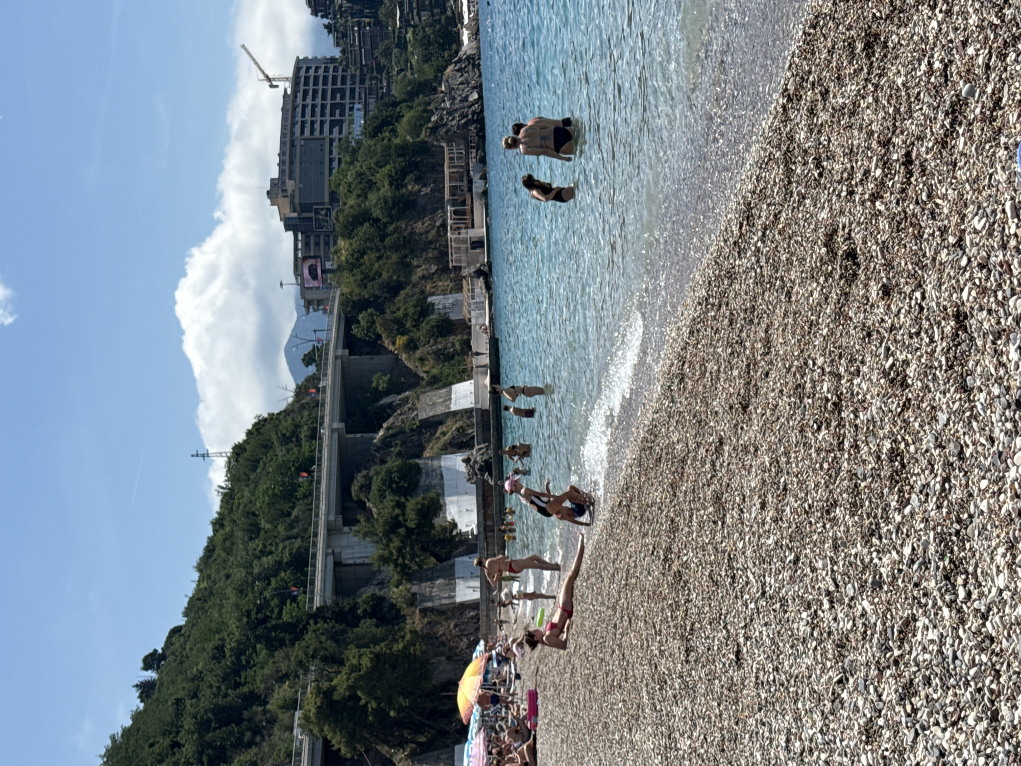 The image depicts a beach scene in Budva, Montenegro. A pebble beach stretches along the right side of the frame, meeting the blue water of the Adriatic Sea. People are swimming and wading in the water, while others sunbathe or relax on the beach. Beach umbrellas and chairs are visible near the bottom of the image. Lush green vegetation covers a hillside adjacent to the beach. In the background, concrete structures and buildings are visible, including a high-rise building under construction and a bridge-like structure. The sky is blue with scattered white clouds.