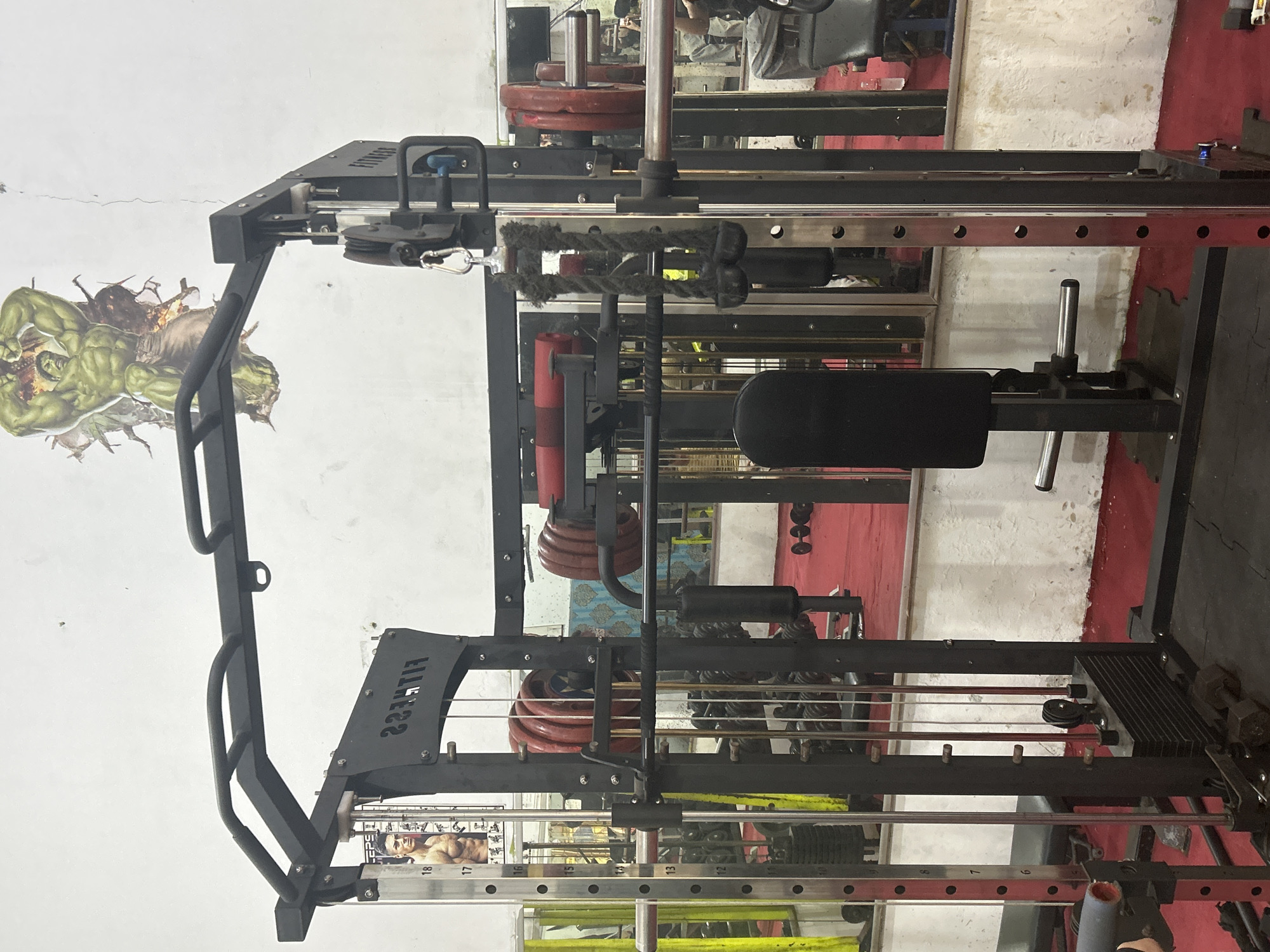 The image depicts a gym in North Jakarta, Indonesia, specifically a Smith machine in the foreground. The machine is black and metallic, with various weight plates, barbells, and a rope attachment visible. Behind the Smith machine, a red floor is prominent, with mirrors reflecting more gym equipment and a person sitting on a bench. The walls are white and cracked, decorated with a poster of The Hulk and another poster featuring a muscular man. The overall scene suggests an active but somewhat dated gym environment.