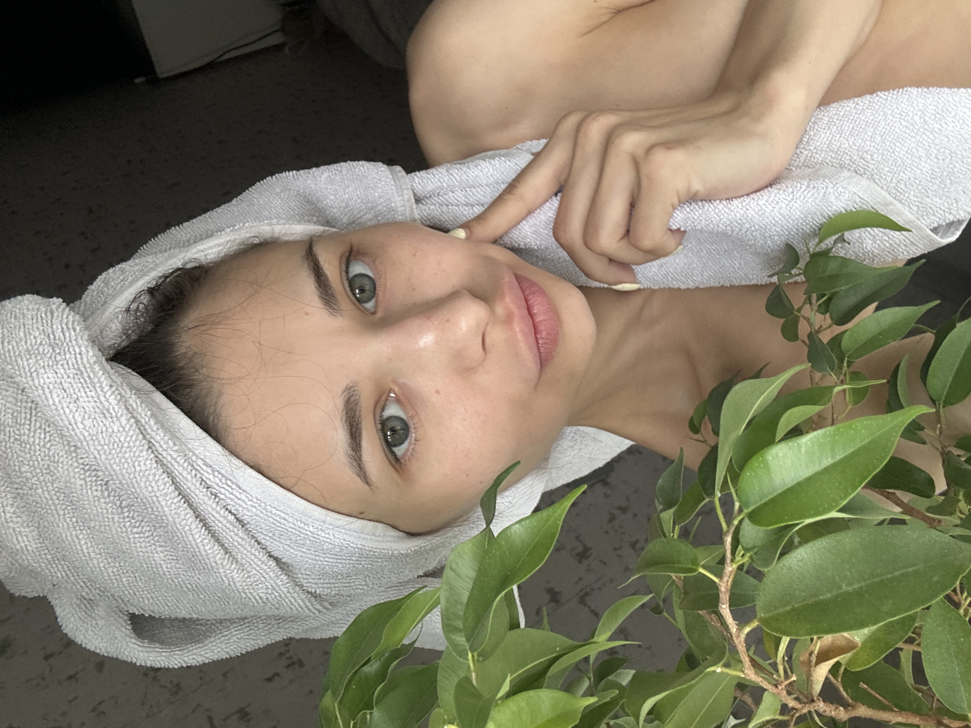 A young woman with fair skin, dark eyebrows, and green eyes lies on what appears to be a gray floor. She has a white towel wrapped around her head. The woman is touching her cheek with her right hand, on which some yellow cream or lotion is visible. A green leafy plant is positioned near her right shoulder. The location of the image is unknown and there is no verifiable information connecting the image to Izhevsk, Russia.